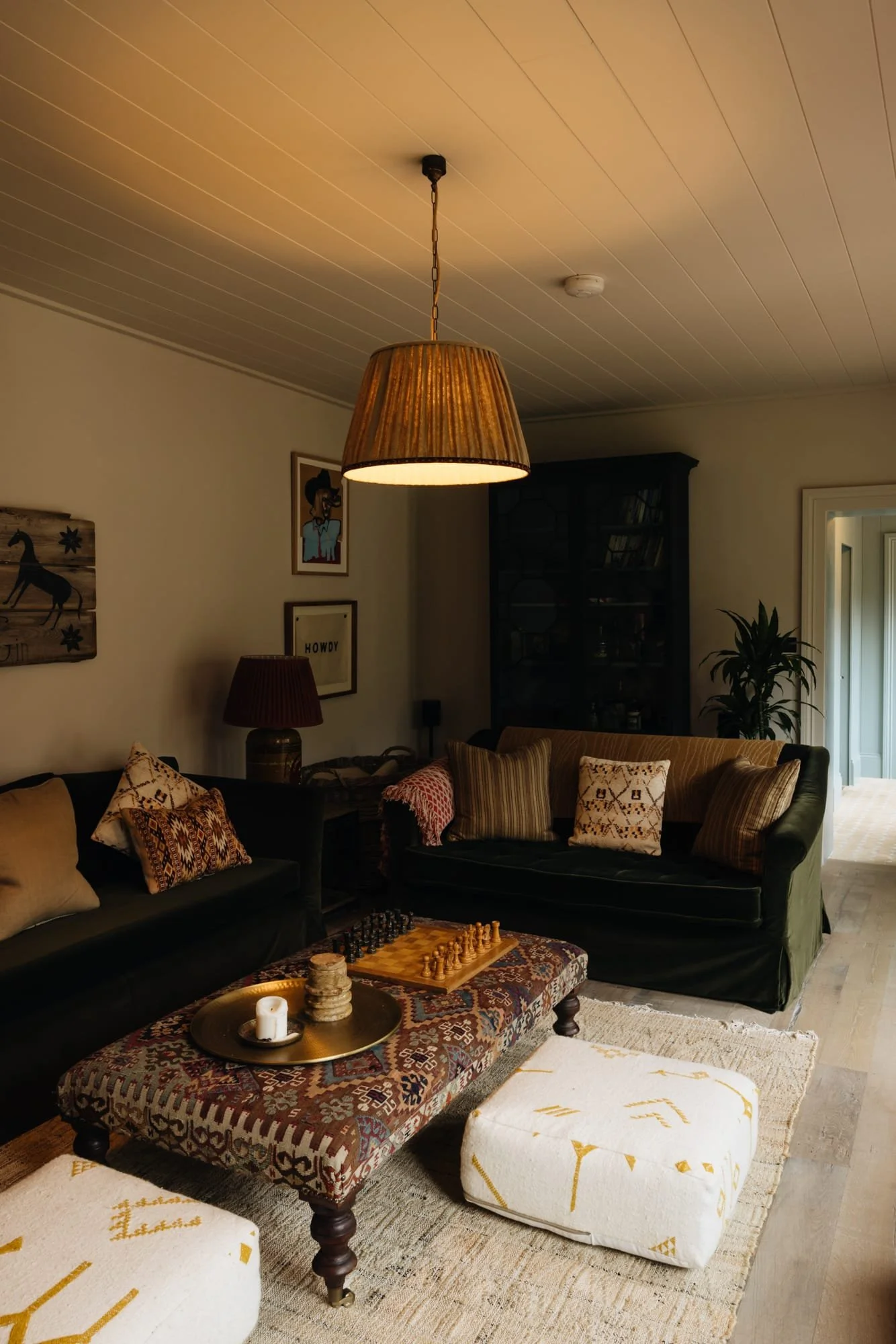 A cozy living room with two dark green sofas, decorative pillows, a patterned ottoman with a chessboard, and a tray with candles and stones. The room is illuminated by a hanging pendant lamp and a table lamp, with wall art and a bookshelf in the back
