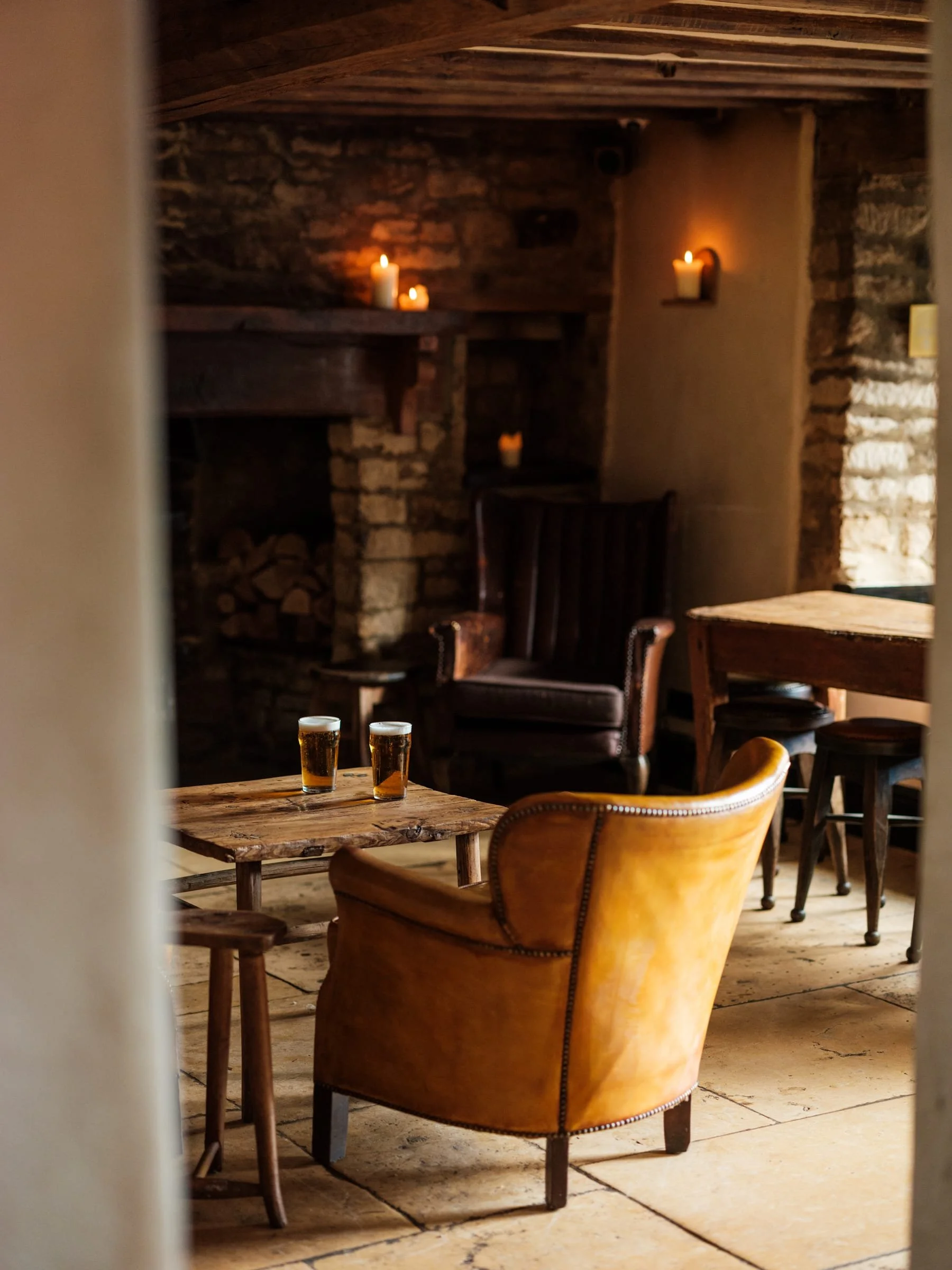 A cozy room with brick fireplace, candles, wooden furniture, and two glasses of beer on a rustic wooden table.