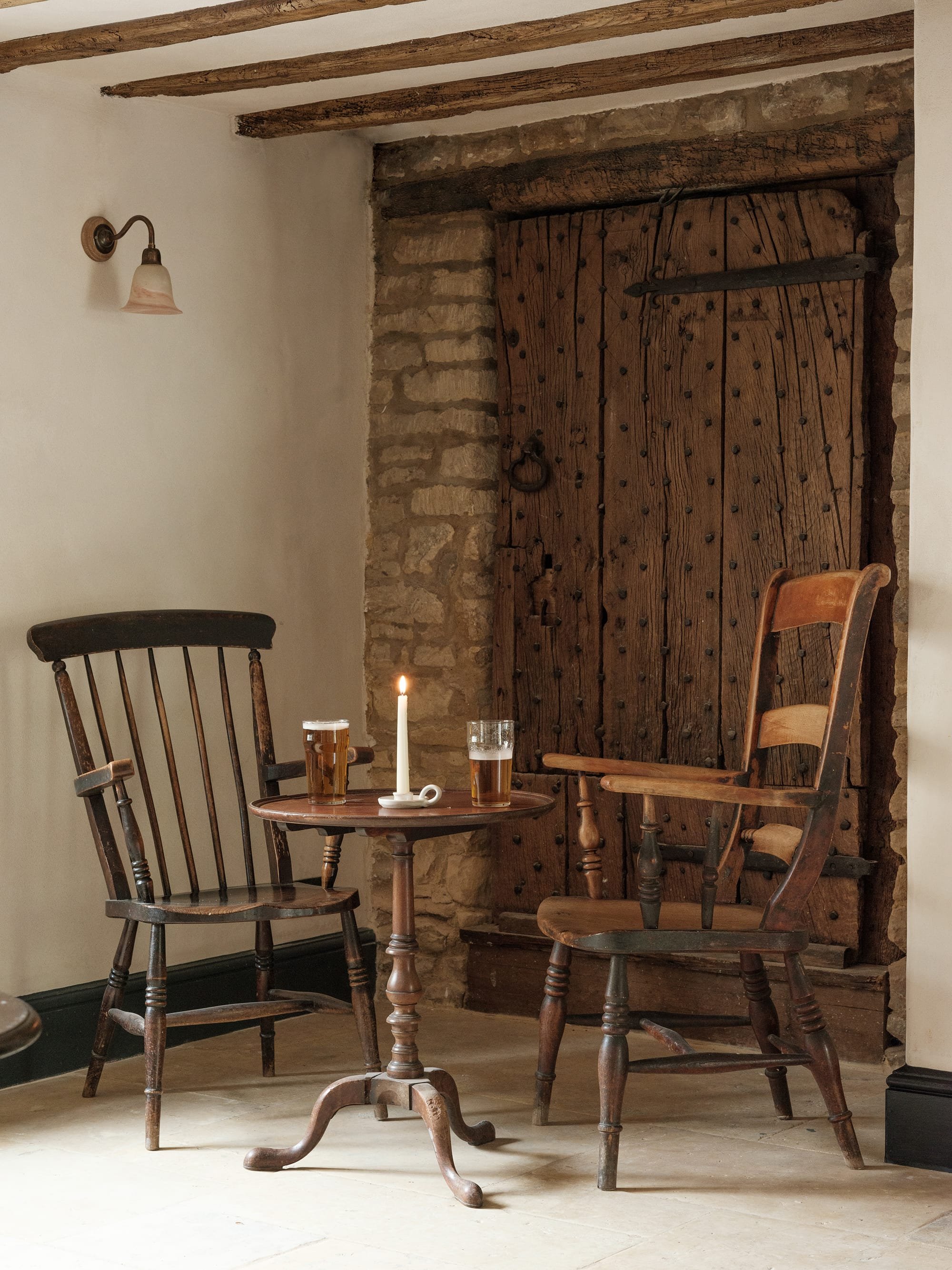 Vintage rustic room with a small round wooden table, two wooden chairs, a lit candle, and two pints of beer. A large wooden door with iron details and stone wall accents is in the background.