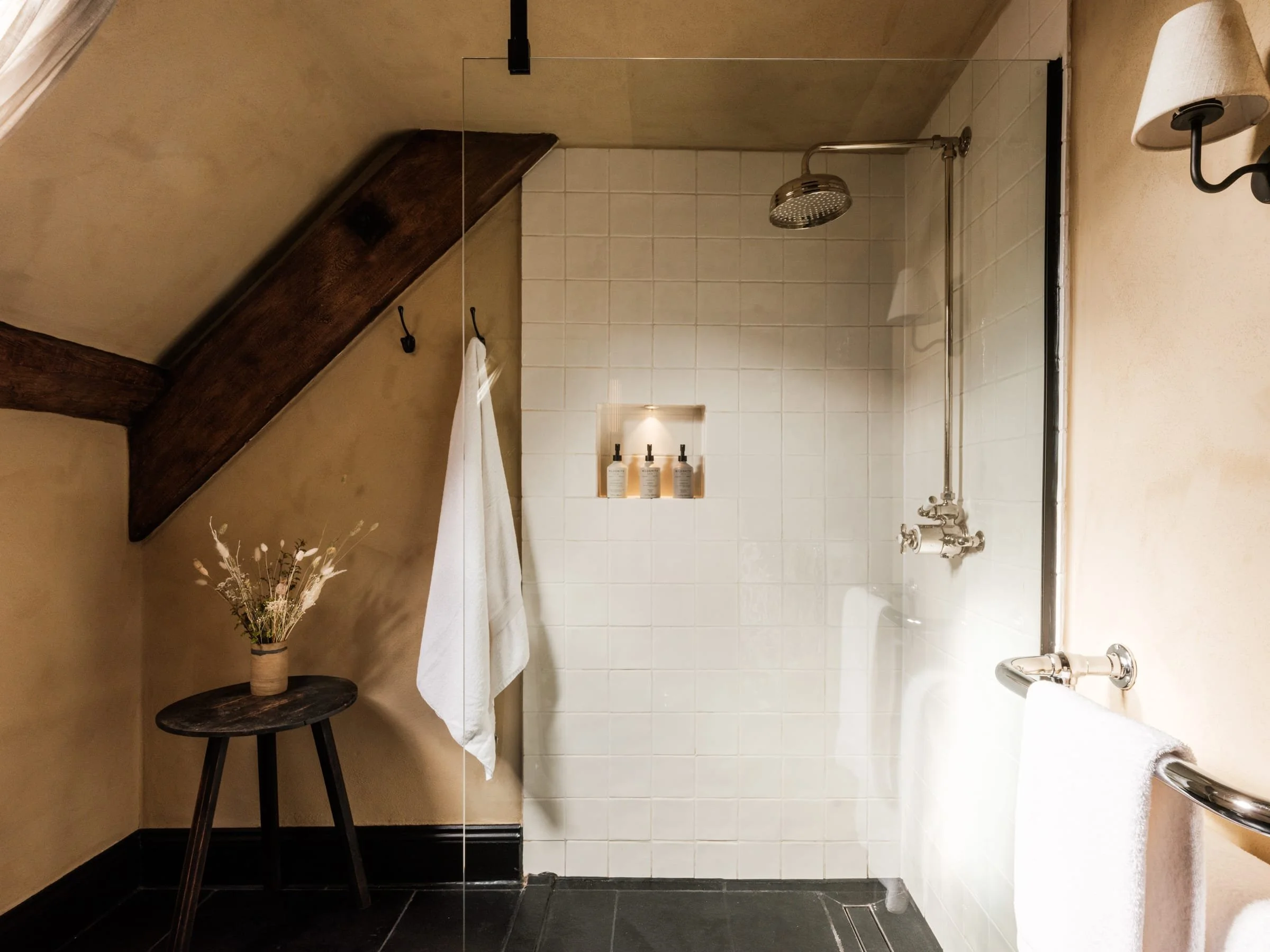 A cozy bathroom with a sloped ceiling, featuring a walk-in shower with white tiled walls, a round rain showerhead, and a built-in shelf with toiletries. There's a small dark wooden stool beside a beige wall, holding a vase with dried flowers, and a w