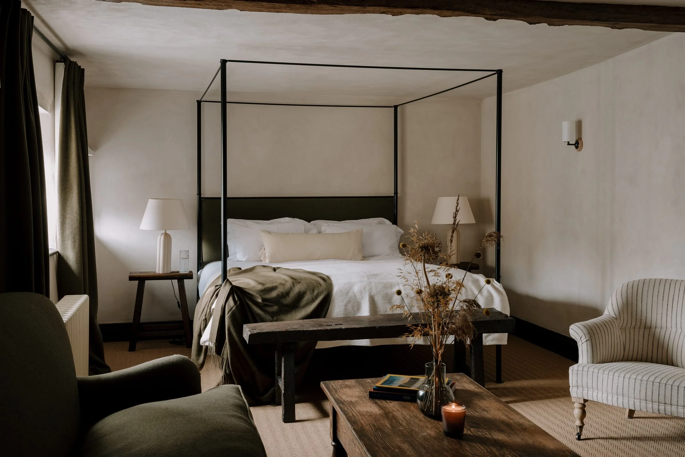 A bedroom with a four-poster bed, two bedside tables with lamps, a coffee table with books and a candle, and sitting furniture including an armchair and a sofa. The room has neutral-colored walls and curtains.