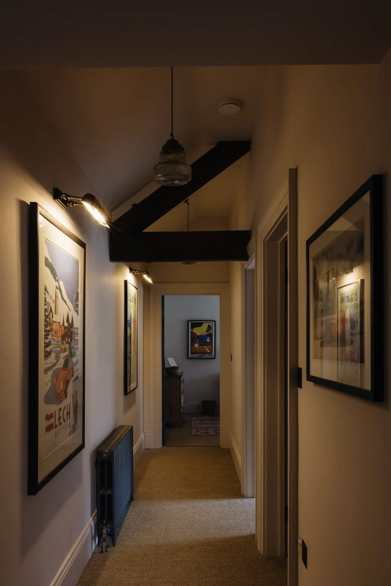 A narrow hallway with framed posters on both walls, a small radiator below the posters on the left side, and a doorway at the end leading to a room with a small cabinet and a framed picture. The hallway has beige carpeting and is lit by wall-mounted 