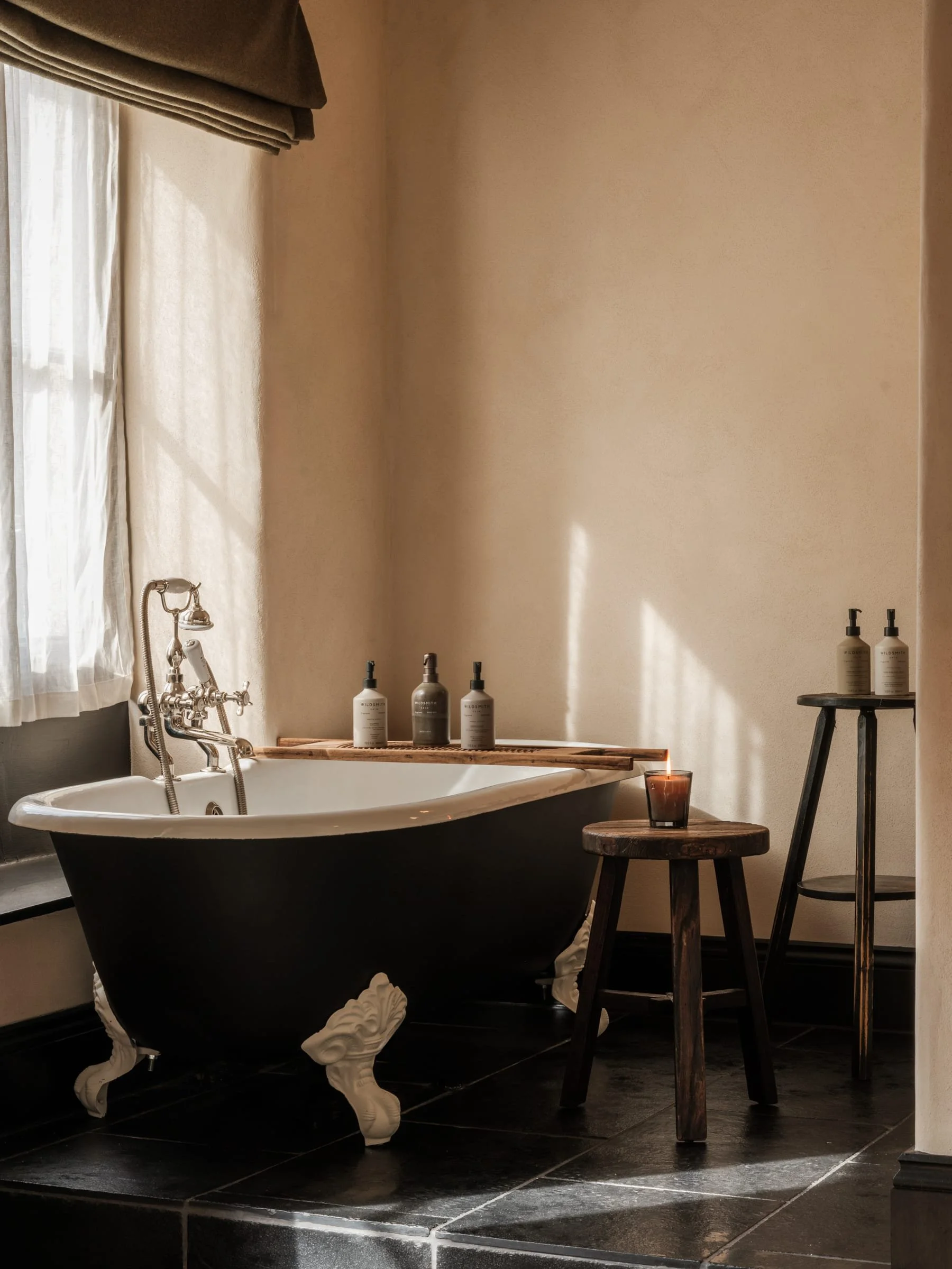 A vintage clawfoot bathtub with black exterior and white interior, placed on a black tiled floor, beside a window with sheer curtains and brown valance. On top of a wooden tray on the tub are three bottles. Near the tub on the floor, there are two sm