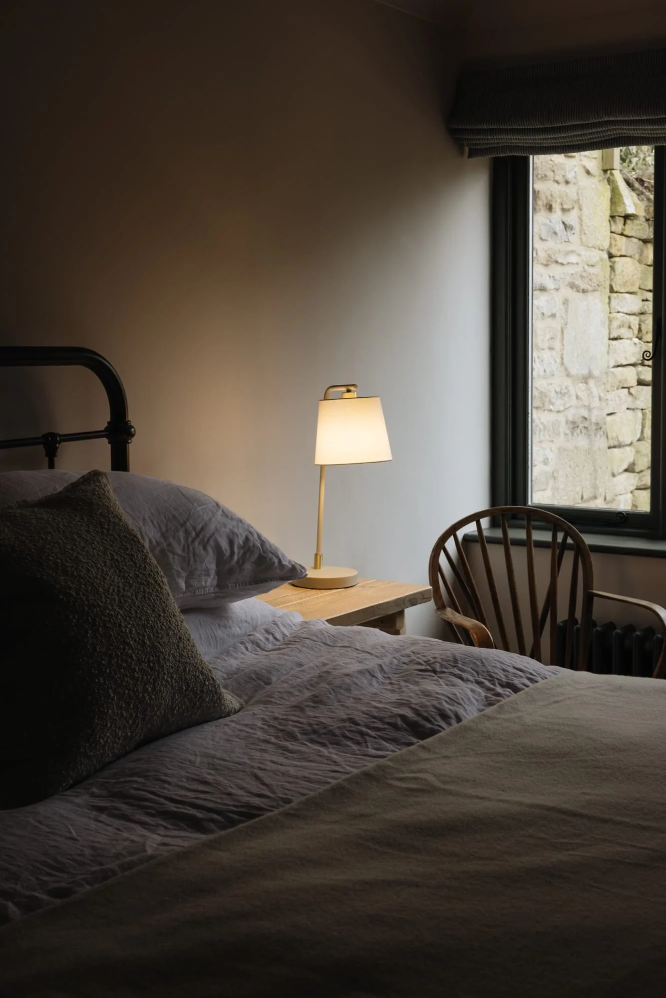 A cozy bedroom with a bed, a bedside lamp, a wooden chair near a window, and a stone wall outside visible through the window.