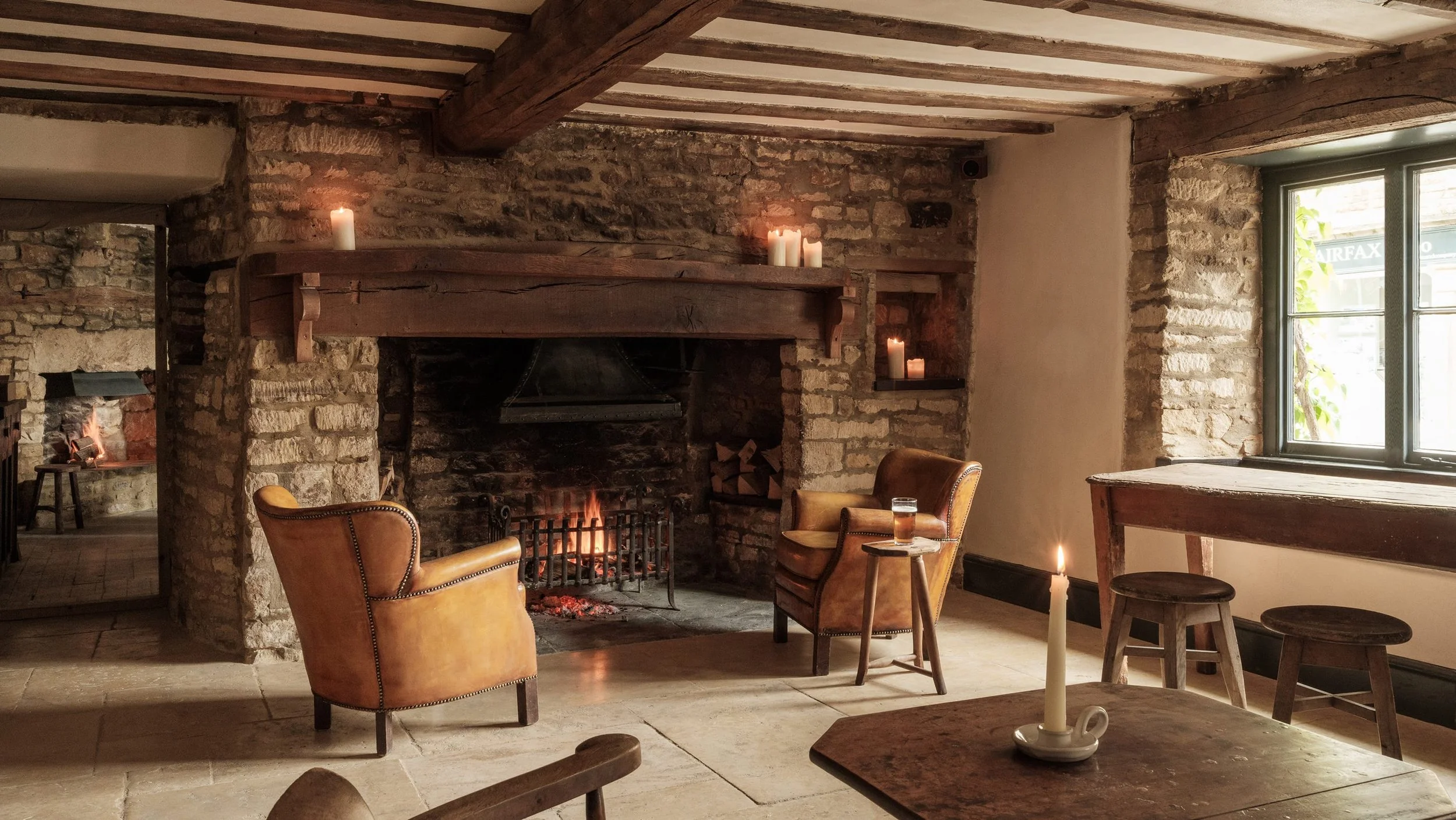 Cozy rustic living room with a lit fireplace, featuring leather armchairs, a wooden table with a candle, and a window letting in natural light, with candles on the mantel and shelf.