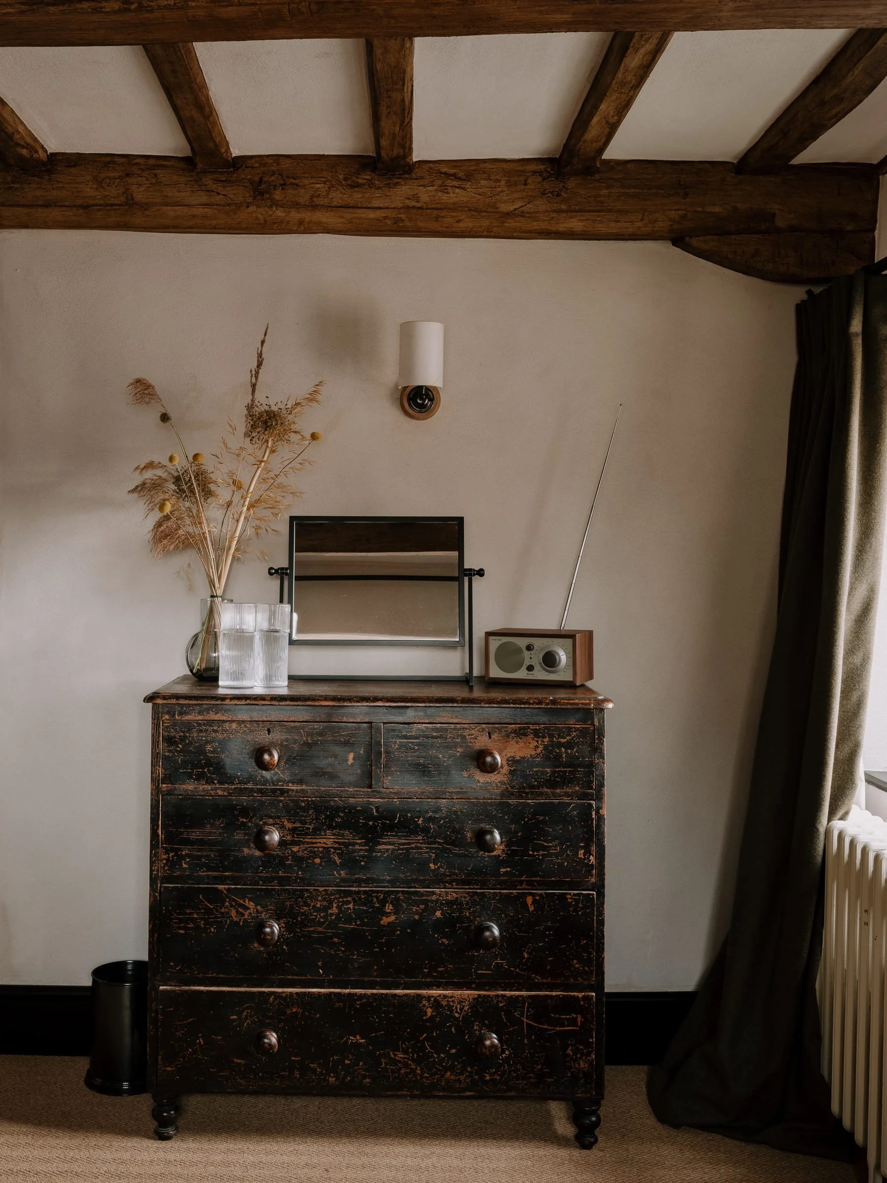 A vintage wooden chest of drawers with a distressed black finish, on a beige carpet near a window with dark curtains. On top of the chest, there is a glass vase with dried pampas grass and other dried plants, a small mirror, a retro radio with an ant