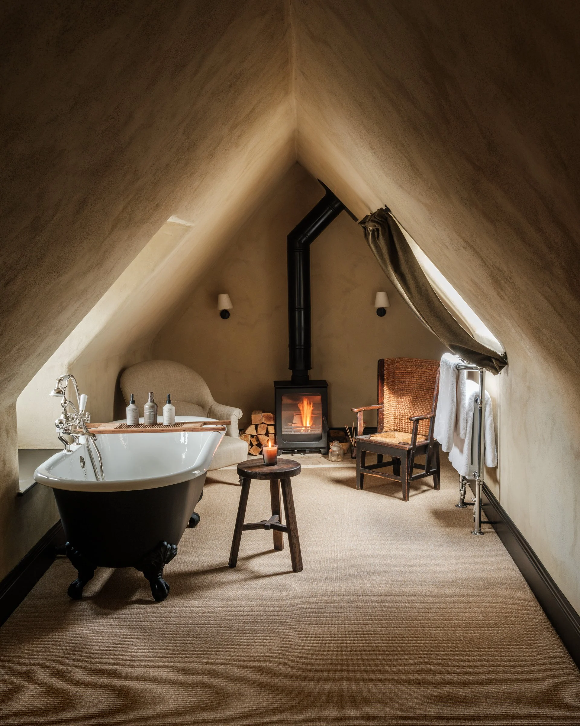 A cozy attic room with a rolled arm sofa, a clawfoot bathtub with toiletries, a wood-burning stove with logs, a chair, a small table with a lit candle, and a slanted window with a curtain.