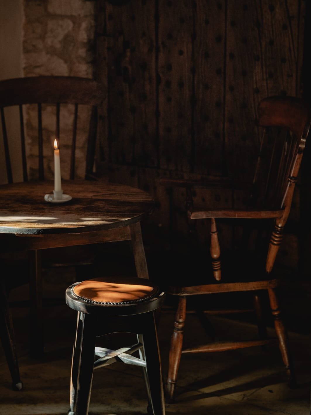 A dark room with a rustic wooden table, a wooden chair, a black stool, and a single lit candle.