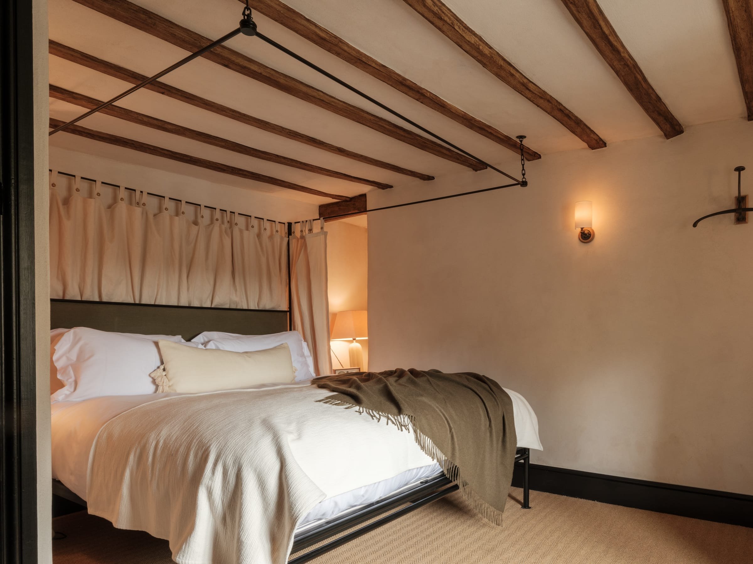 A cozy bedroom with a large bed draped in white bedding and a beige throw blanket. The bed features decorative pillows and a canopy with curtains suspended from a ceiling-mounted rod. The room has a rustic ceiling with wooden beams, a wall sconce, an