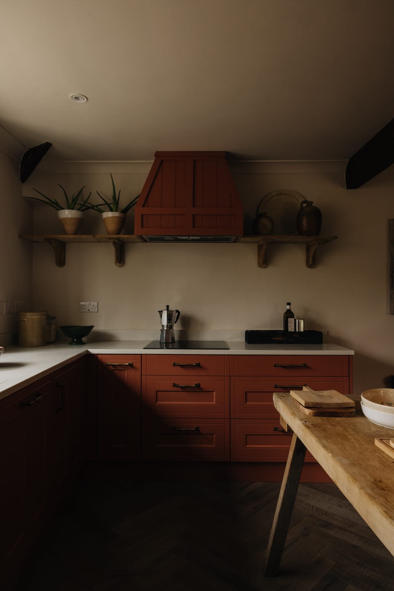 Modern kitchen with red cabinets, wooden open shelves holding potted plants and decorative items, a white countertop with a coffee maker, and a wooden table with bowls and cutting boards.
