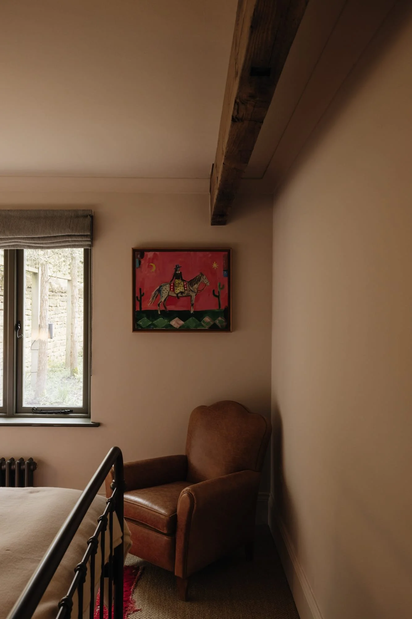 A corner of a bedroom showing a brown upholstered armchair, a window with beige blinds, a colorful painting of a cowboy on a horse with cacti on the wall, and part of a bed with black metal frame.