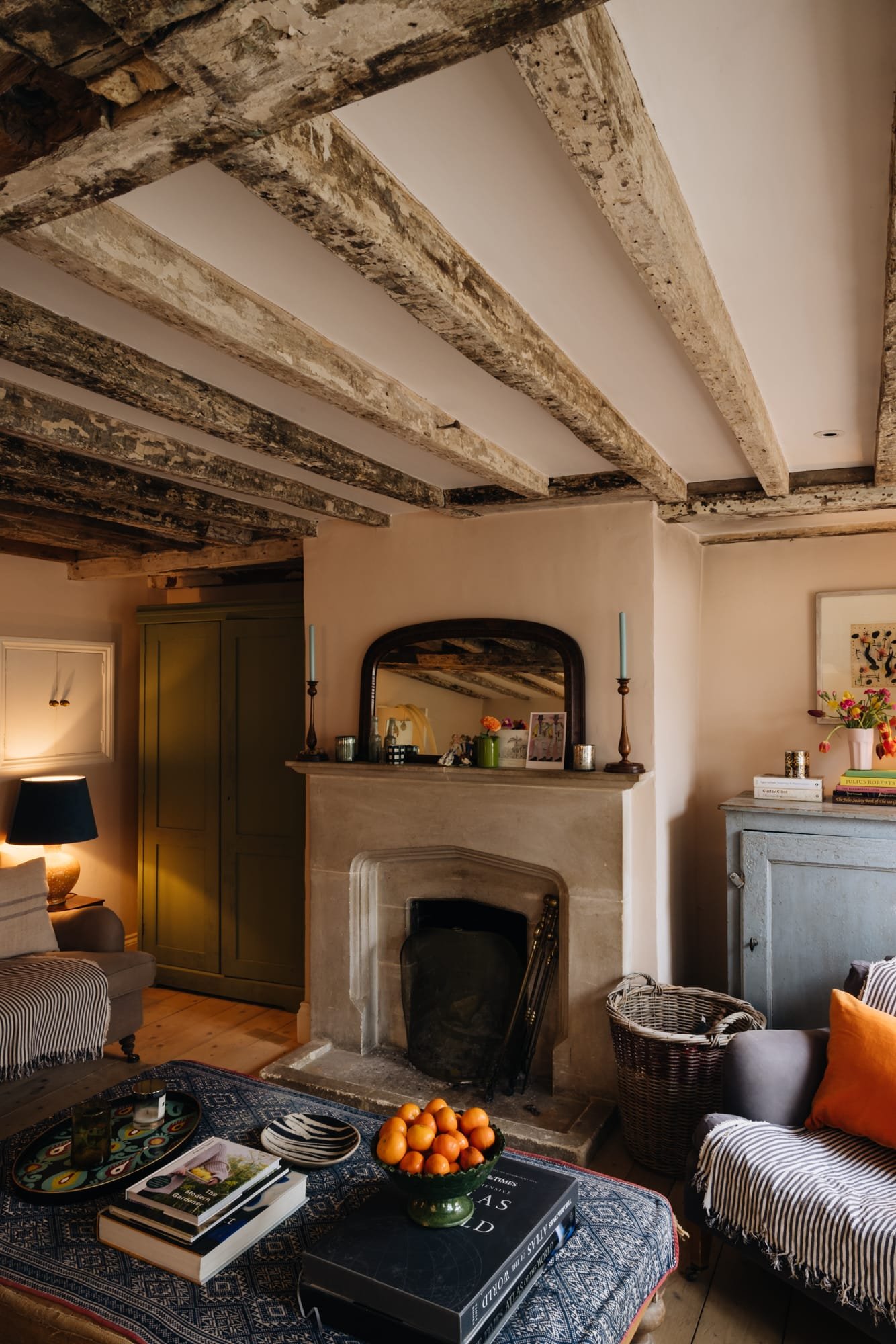 Cozy living room with exposed wooden ceiling beams, a fireplace, and a mix of vintage and modern decor, including books, candles, and a bowl of oranges on a patterned table.