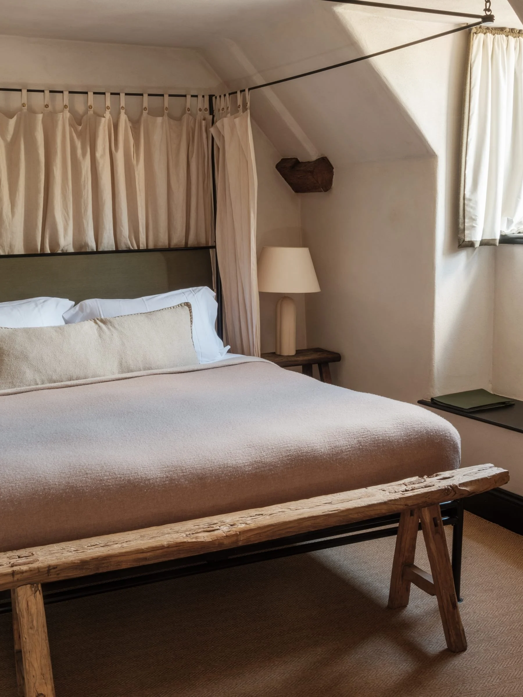 A cozy bedroom with a bed, beige bedding, and a rustic wooden bench at the foot of the bed. There is a beige curtain, a window, and a nightstand with a beige lamp. The room has a sloped ceiling with a small wooden beam.