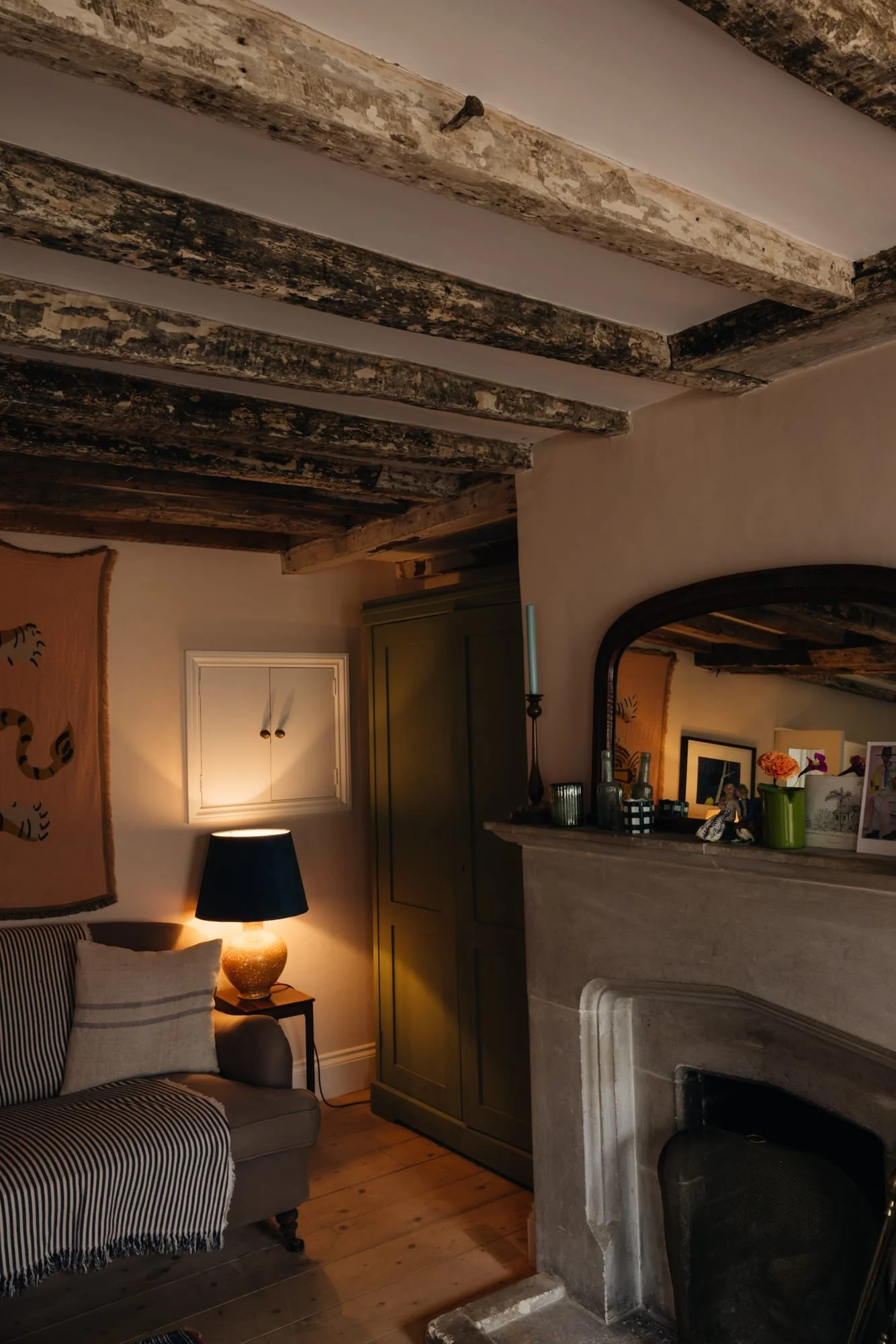 Cozy living room corner with a striped armchair, illuminated by a table lamp with a black shade, a green wardrobe, and a fireplace mantel decorated with candles, vases, and framed photos. Exposed wooden beams on the ceiling and a tapestry on the wall