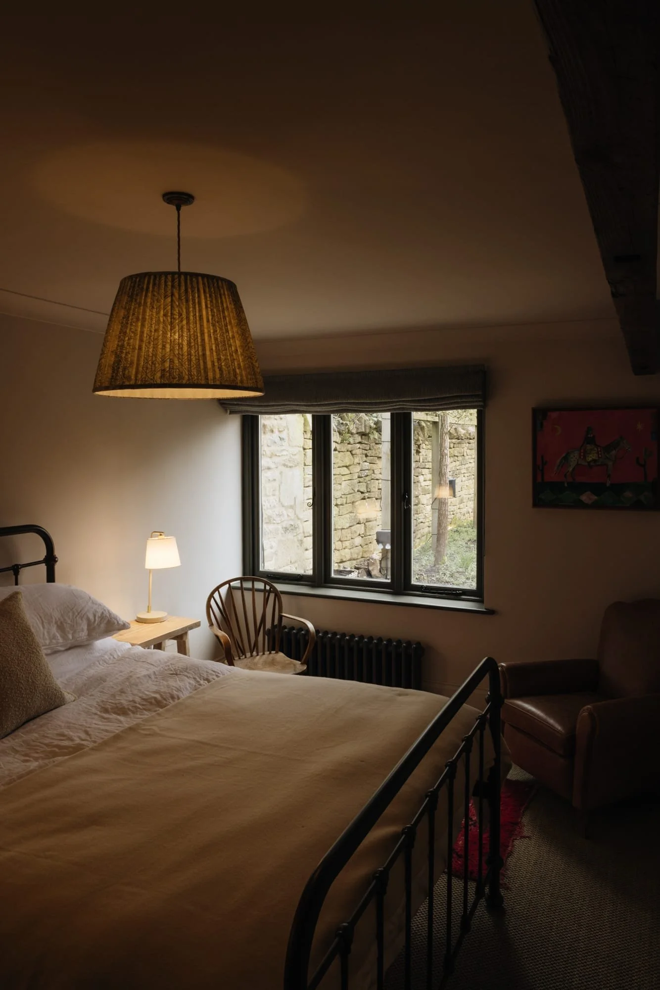 A cozy bedroom with a bed, bedside lamp, wooden chair, armchair, and window looking out onto a stone wall outside.