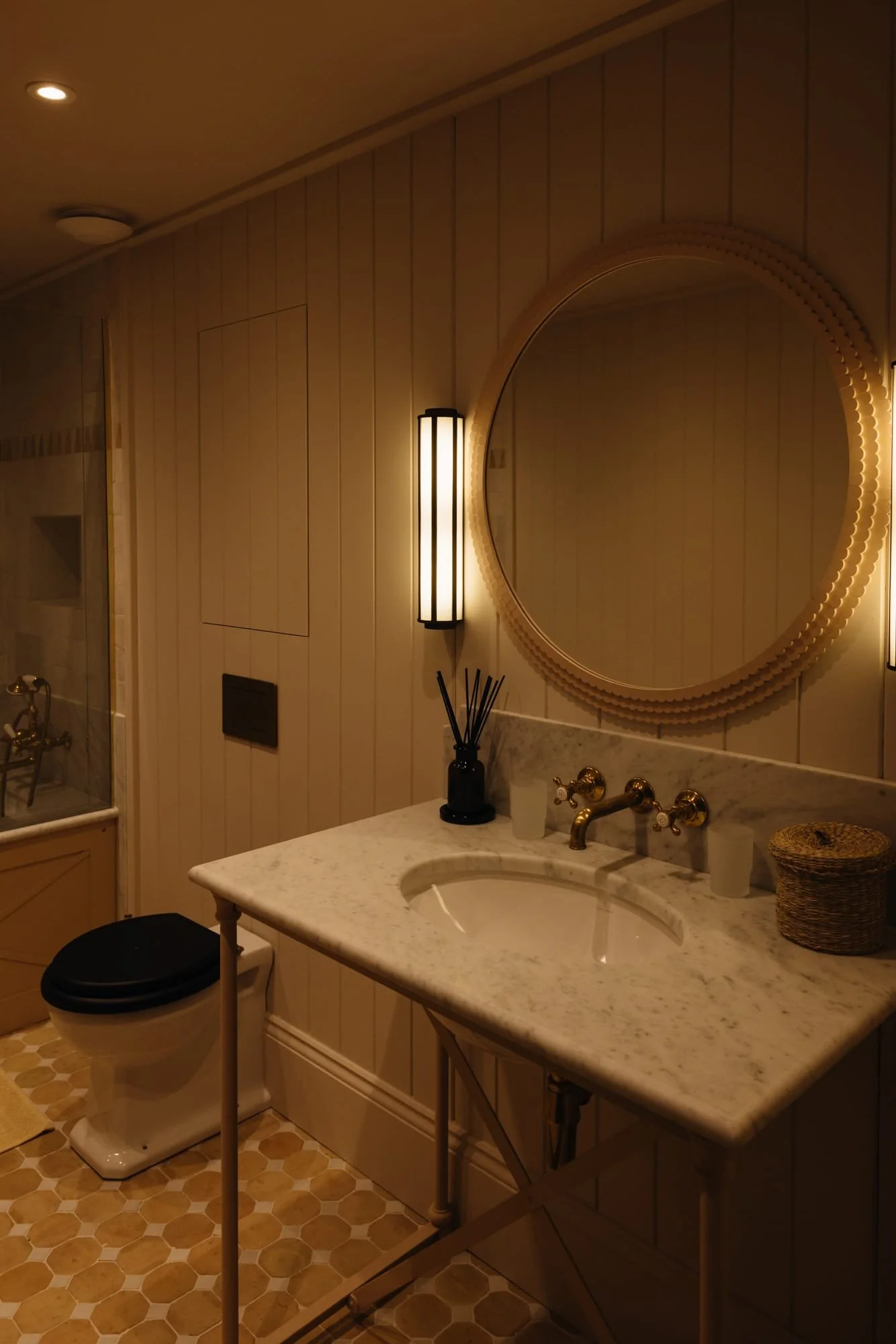 A bathroom with beige paneled walls, a white marble countertop sink, and warm lighting. There is a round mirror, two vertical wall sconces, a black reed diffuser, a basket on the counter, and a toilet with a black seat.