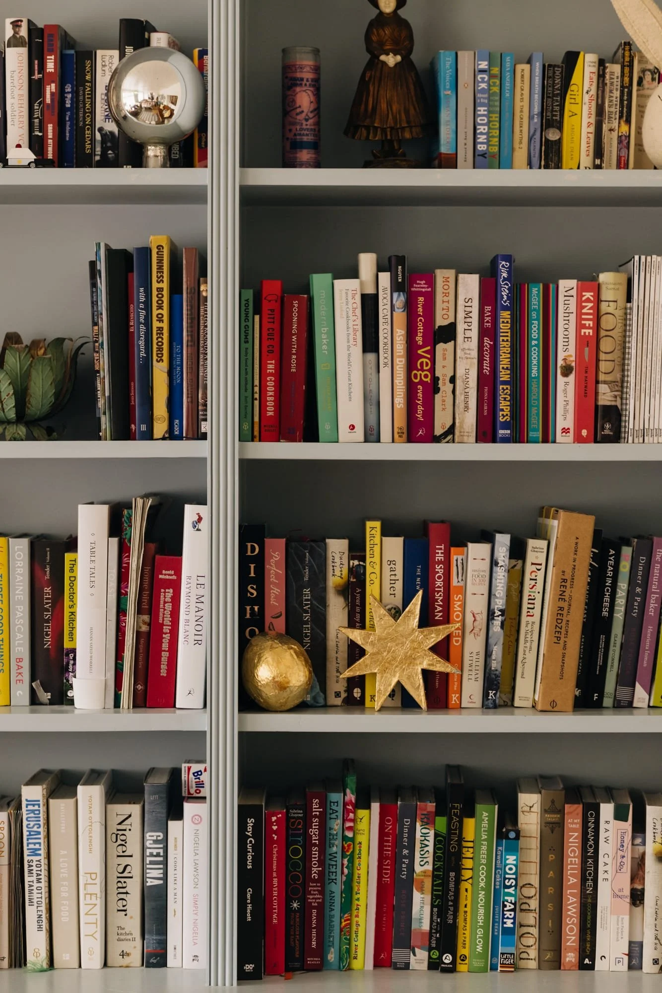 Bookshelves filled with cookbooks, novels, and decorative items, including a gold paper mache globe, a gold star, and a wooden figurine of a woman.