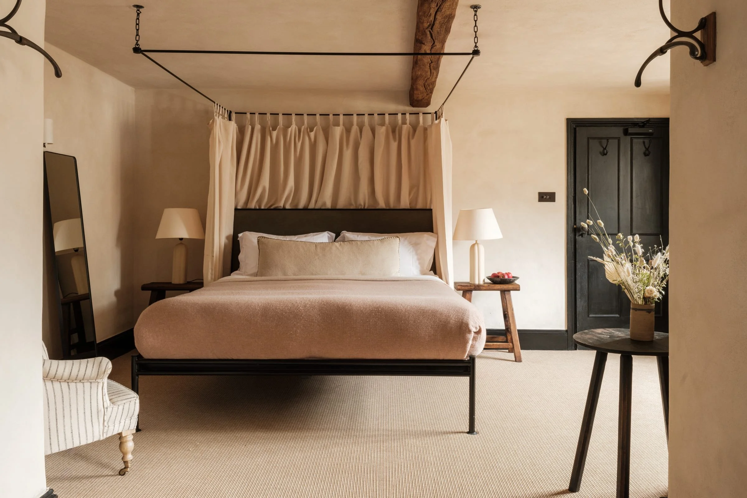 A cozy bedroom with a bed, curtains, two bedside lamps, and a mirror, featuring a vase with flowers and a black door.