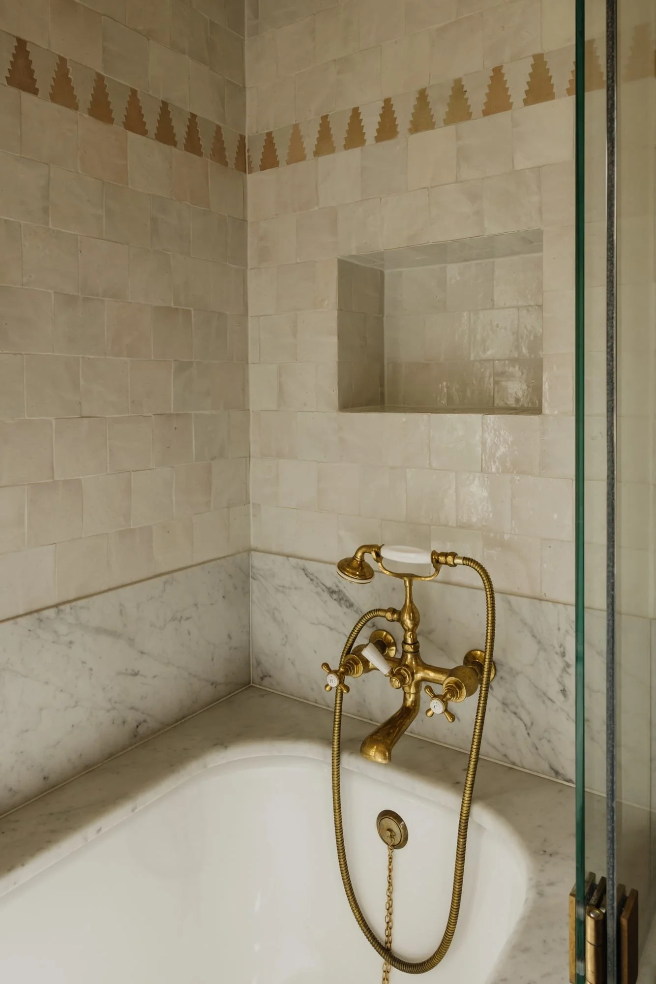 Bathroom with a white bathtub, gold faucet, marble countertop, beige tiled walls, and a built-in wall shelf.