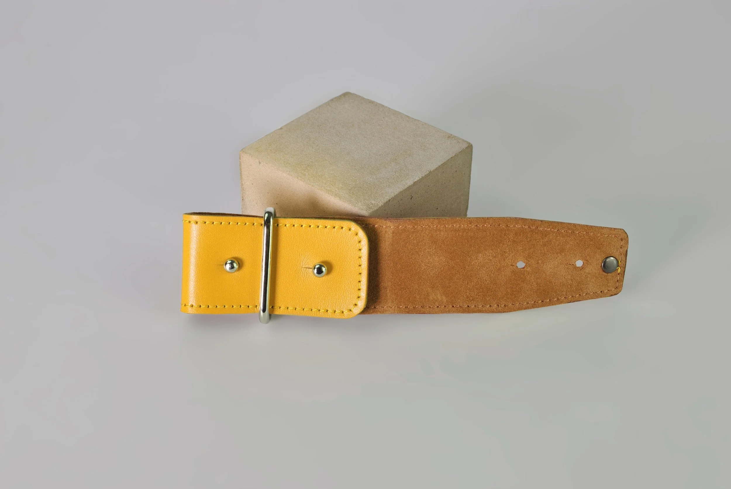 A yellow leather belt with metal buckle, partially unfastened, lying on a gray surface with a beige concrete cube in the background.