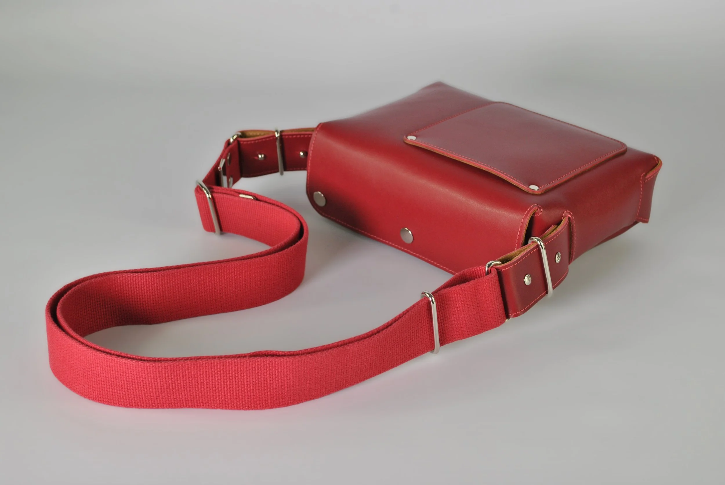Red leather crossbody bag with a wide red fabric strap on a light grey background.