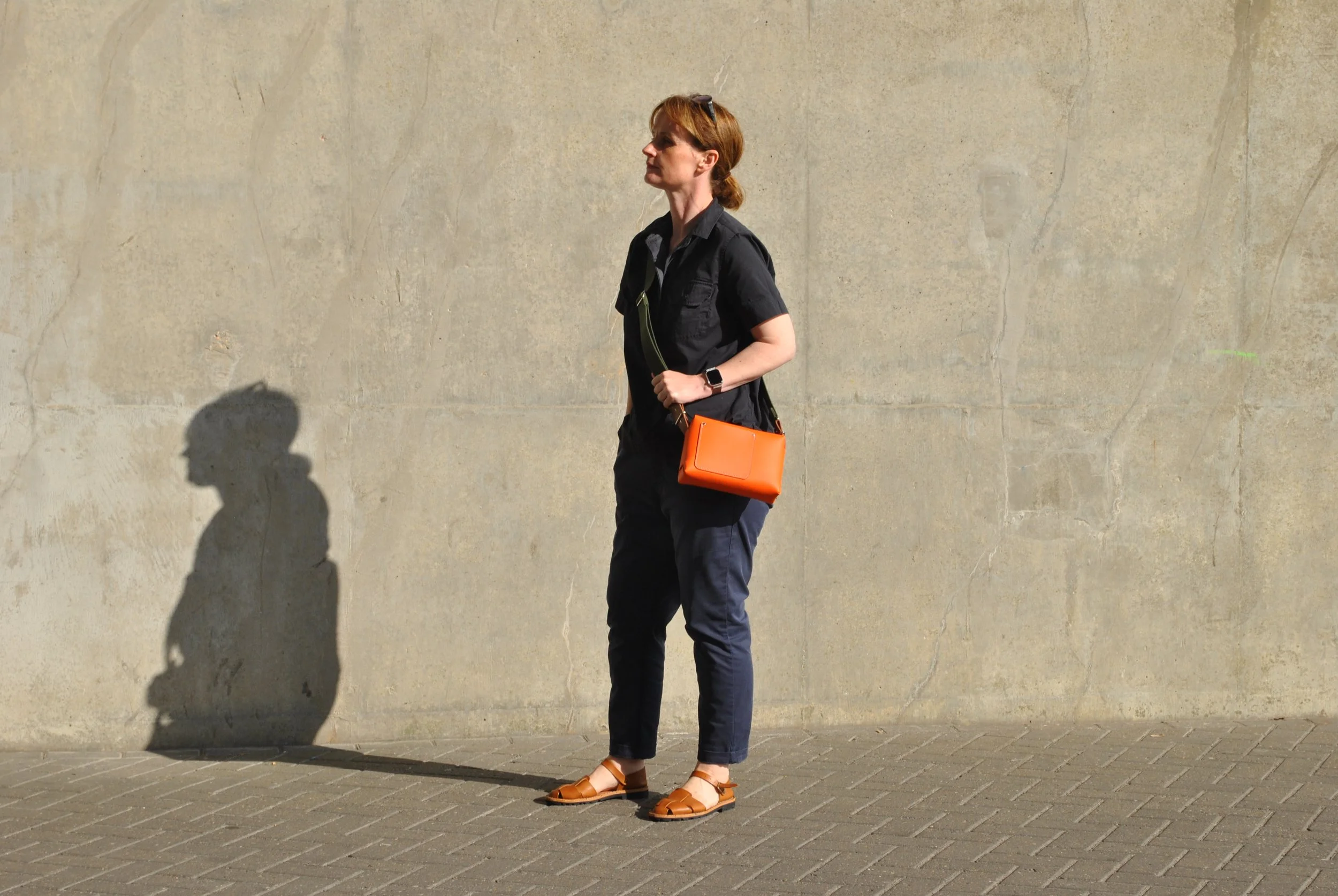 A woman with red hair wearing a black shirt, navy pants, and brown sandals, standing against a beige wall, holding an orange bag. Her shadow is cast on the wall.
