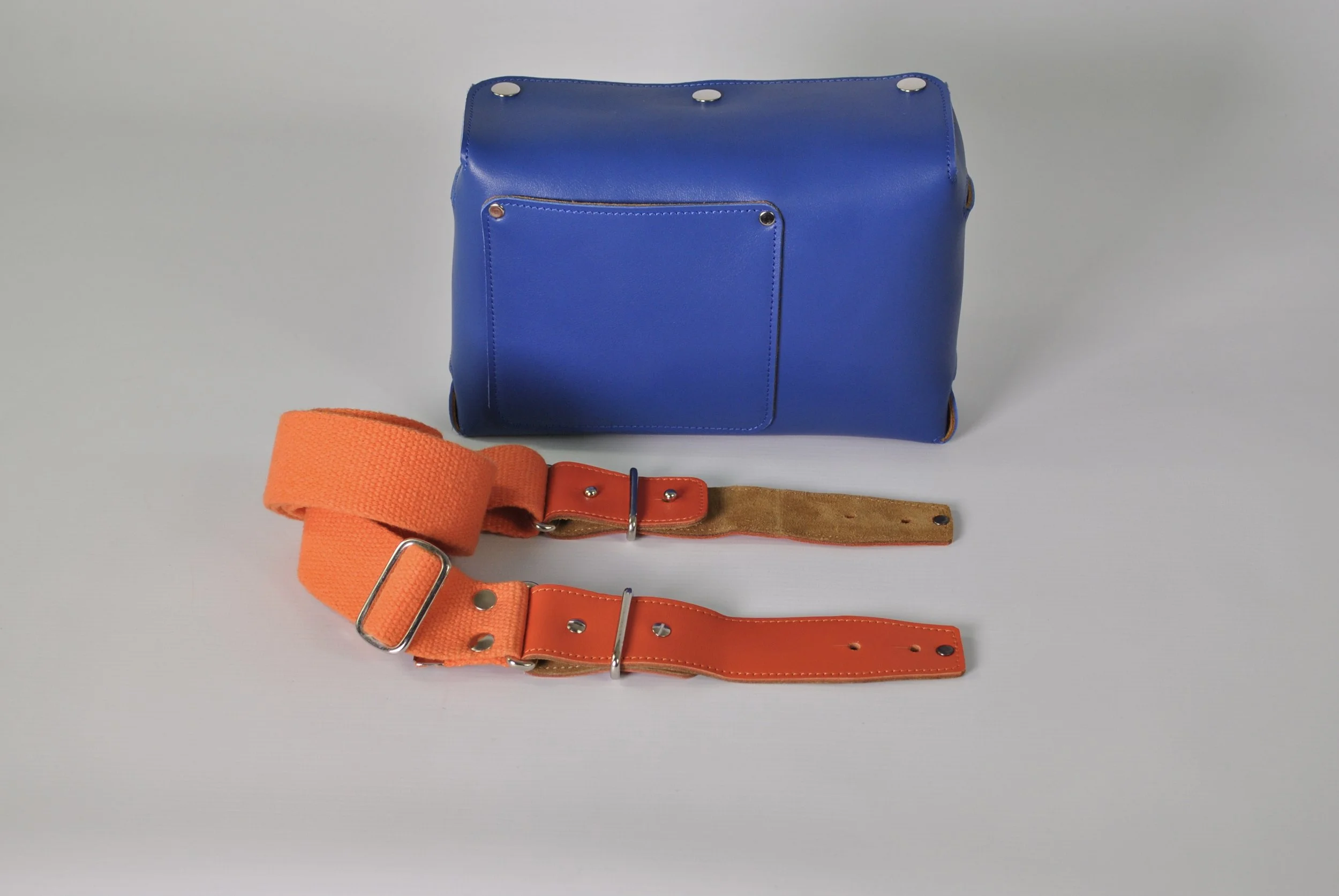 A blue leather bag with a front pocket and metal snaps, placed on a light gray surface. In front of the bag are two belts, one orange with metal loops and a buckle, and one brown suede with an orange leather section and metal hardware.