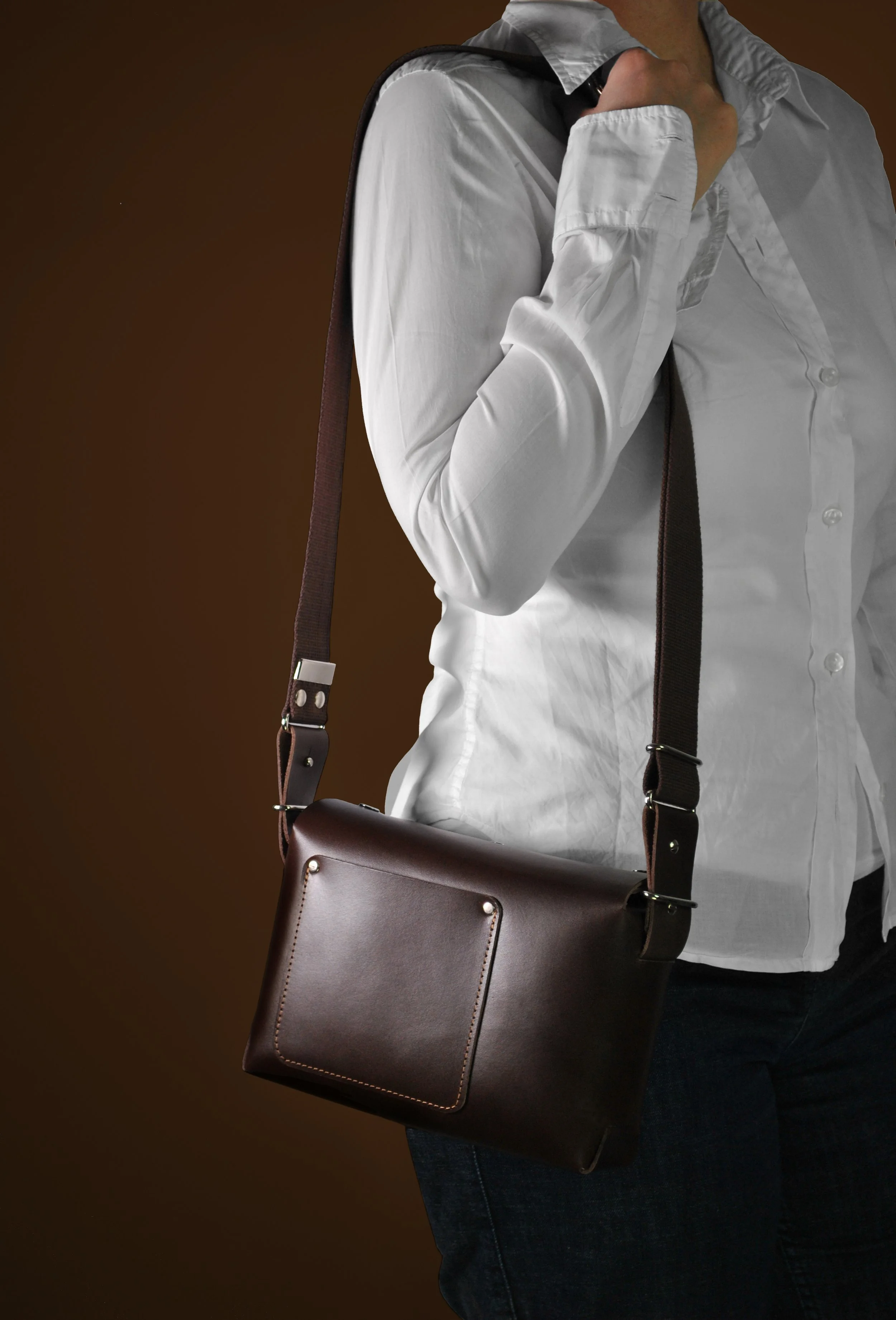 A person wearing a white shirt carrying a dark brown shoulder bag against a brown background.