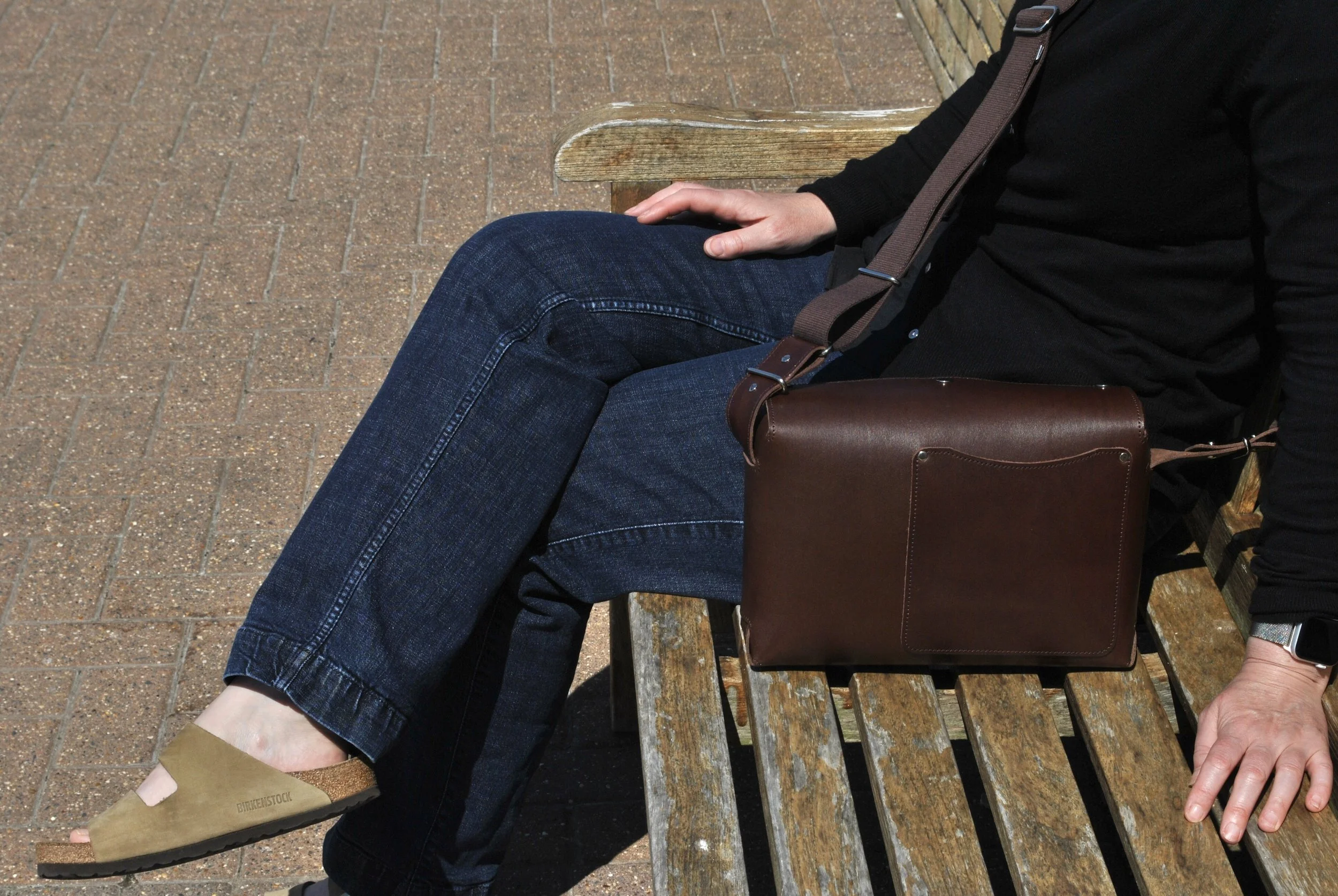 Person sitting on a wooden park bench, wearing blue jeans and beige Birkenstock sandals, with a brown leather bag on their lap. The person is wearing a black long-sleeve shirt and a black smartwatch.