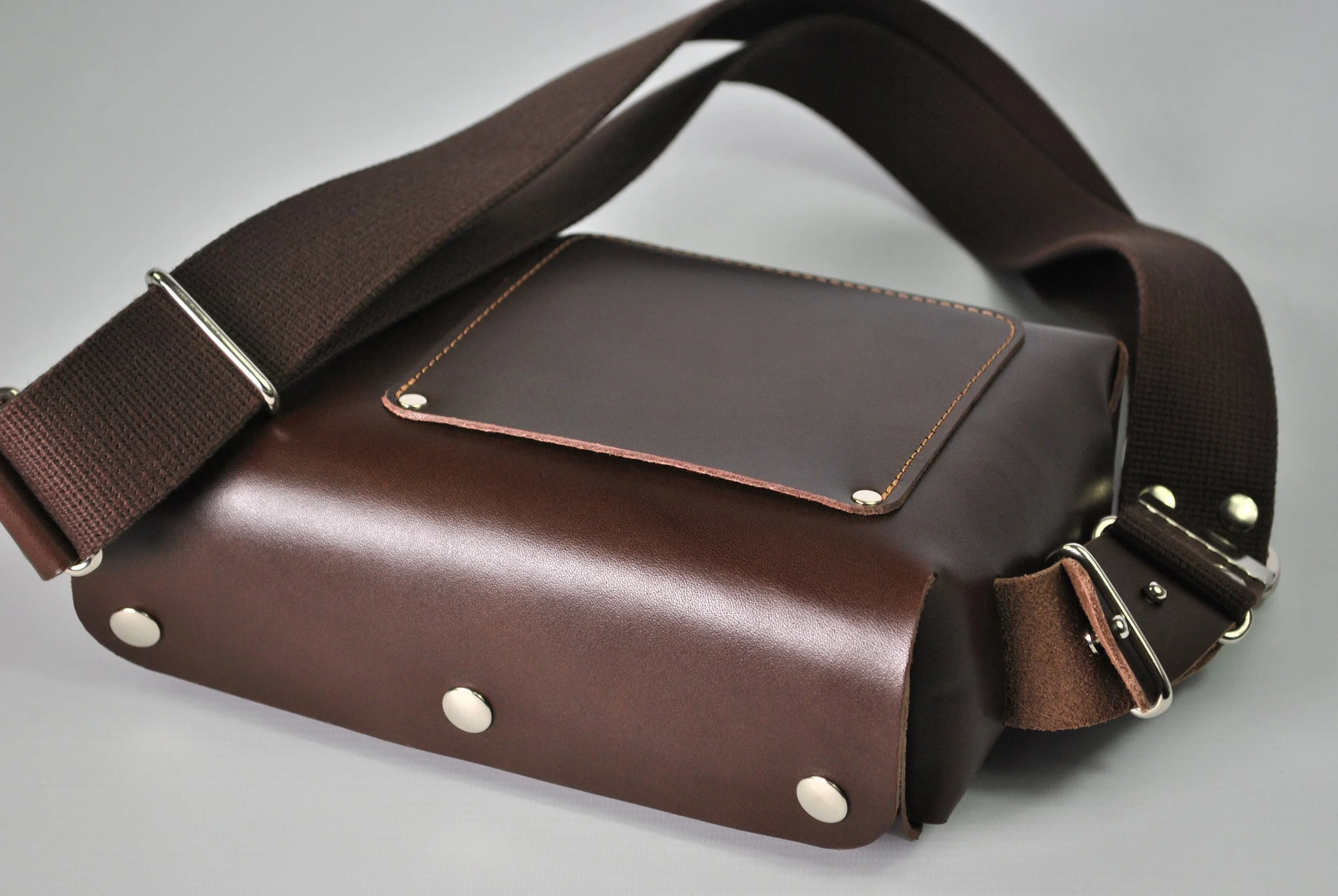 Brown leather shoulder bag with a dark brown front pocket, metal studs on the bottom, and a wide adjustable dark brown strap.