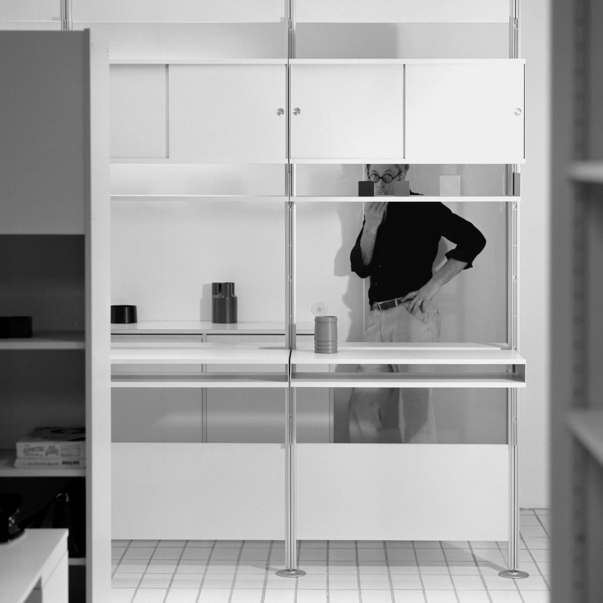 Black and white photo of a person, standing in front of a shelving unit in a room with tiled floor.