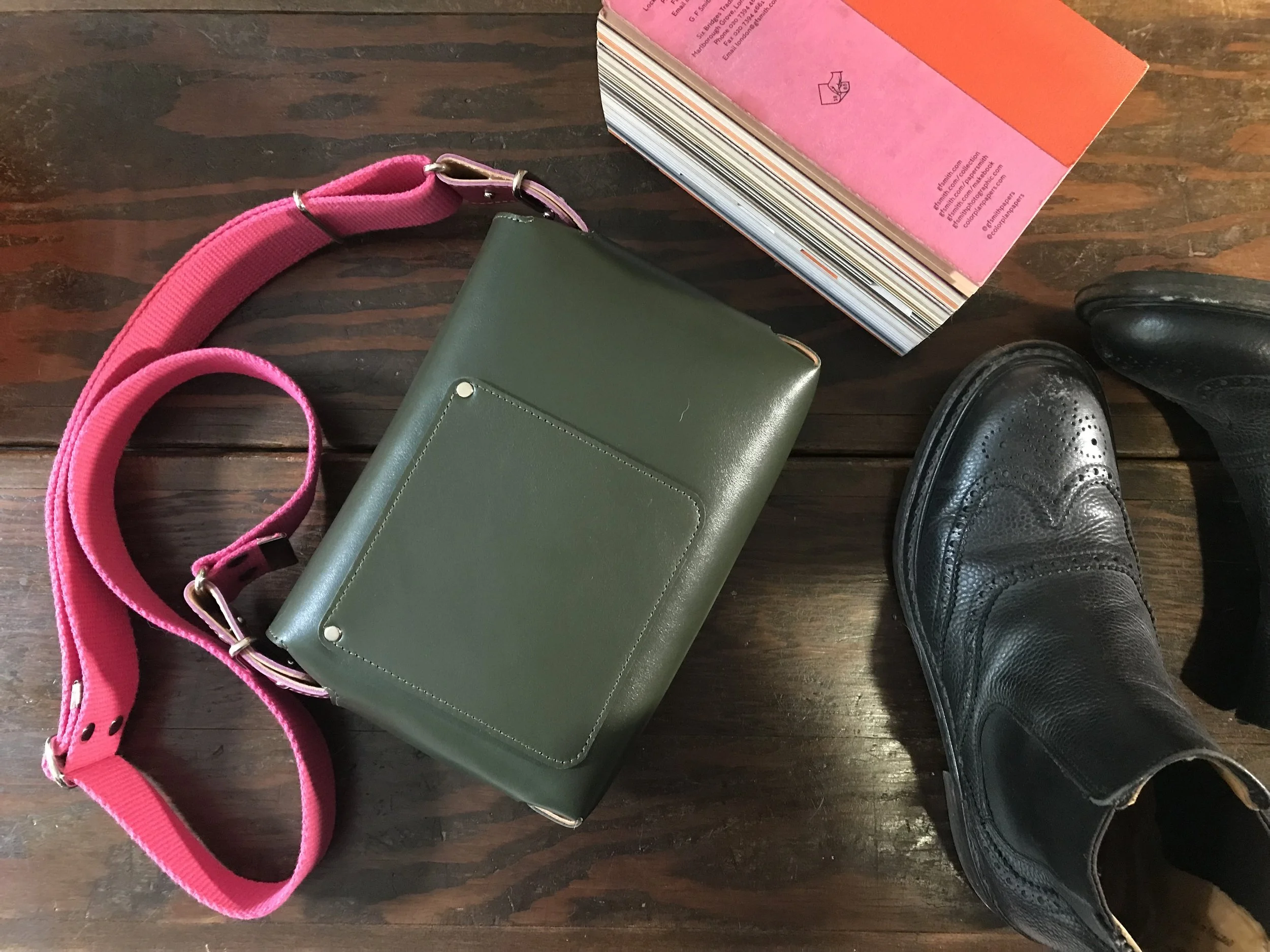 A green handbag with a pink strap, a pair of black dress shoes, and a pink book on a wooden surface.
