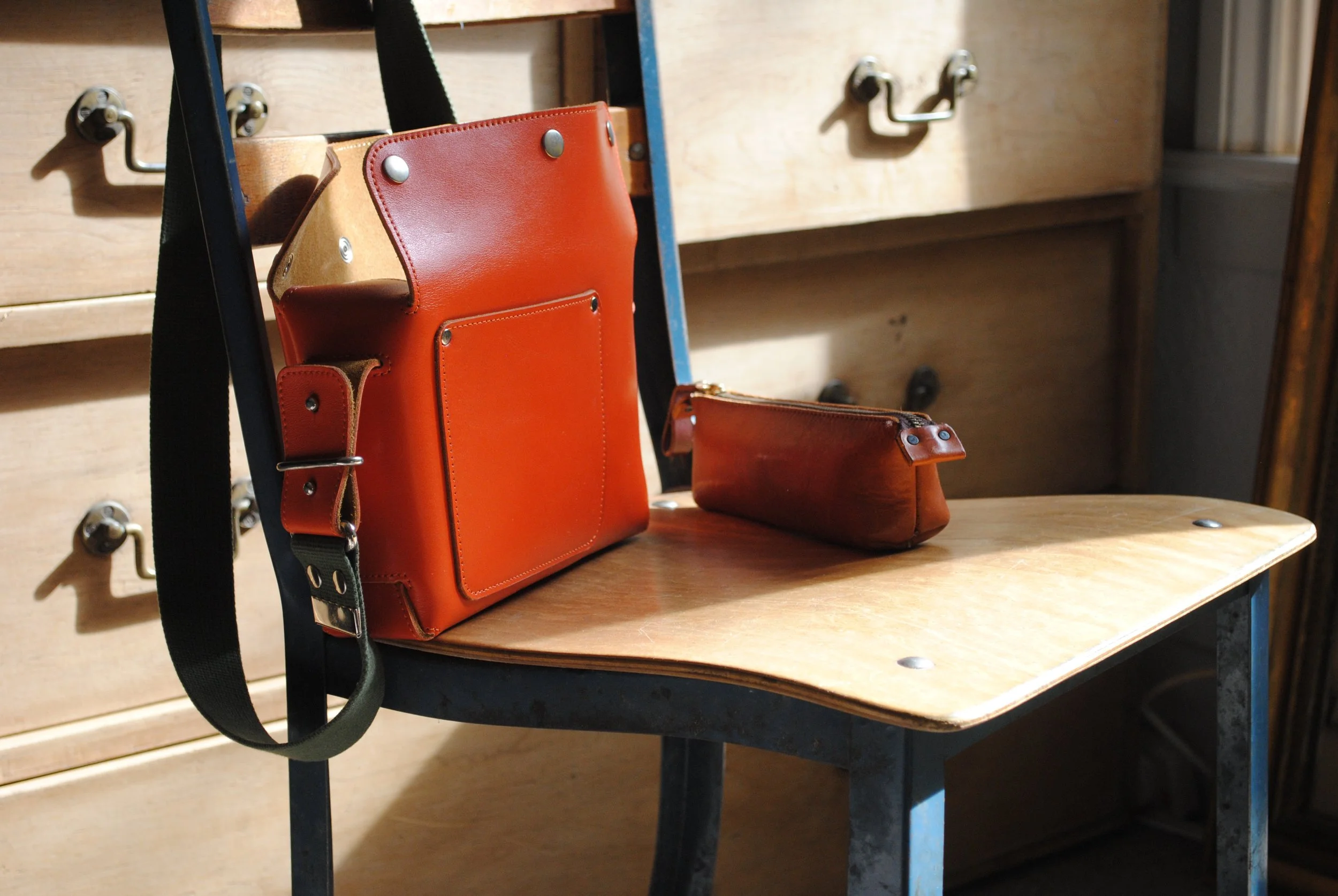 Orange leather shoulder bag and matching pouch on wooden chair in a room with wooden drawers.