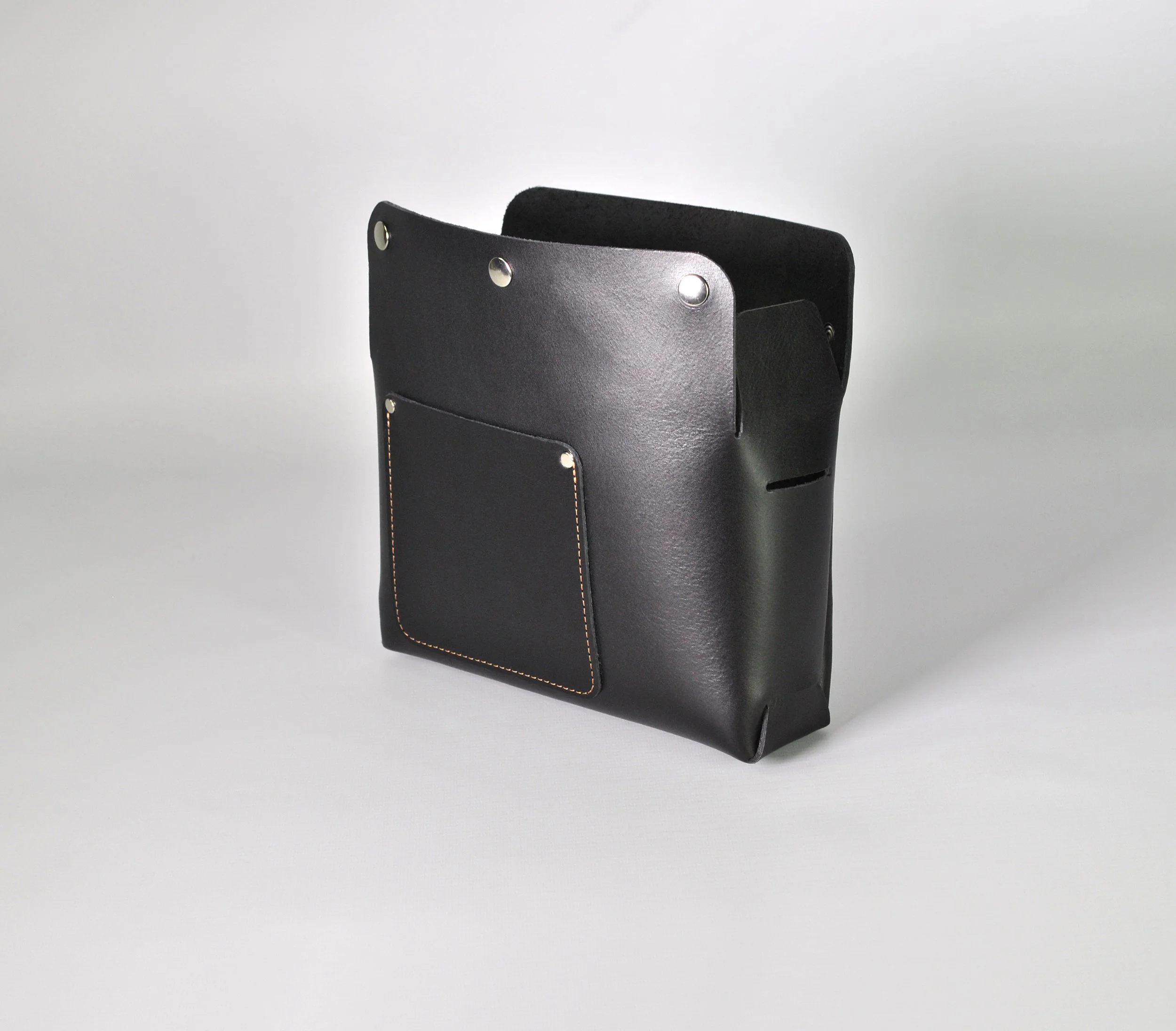 A black leather tote bag with a front pocket and metal rivets on the top corners, placed on a plain, light gray background.