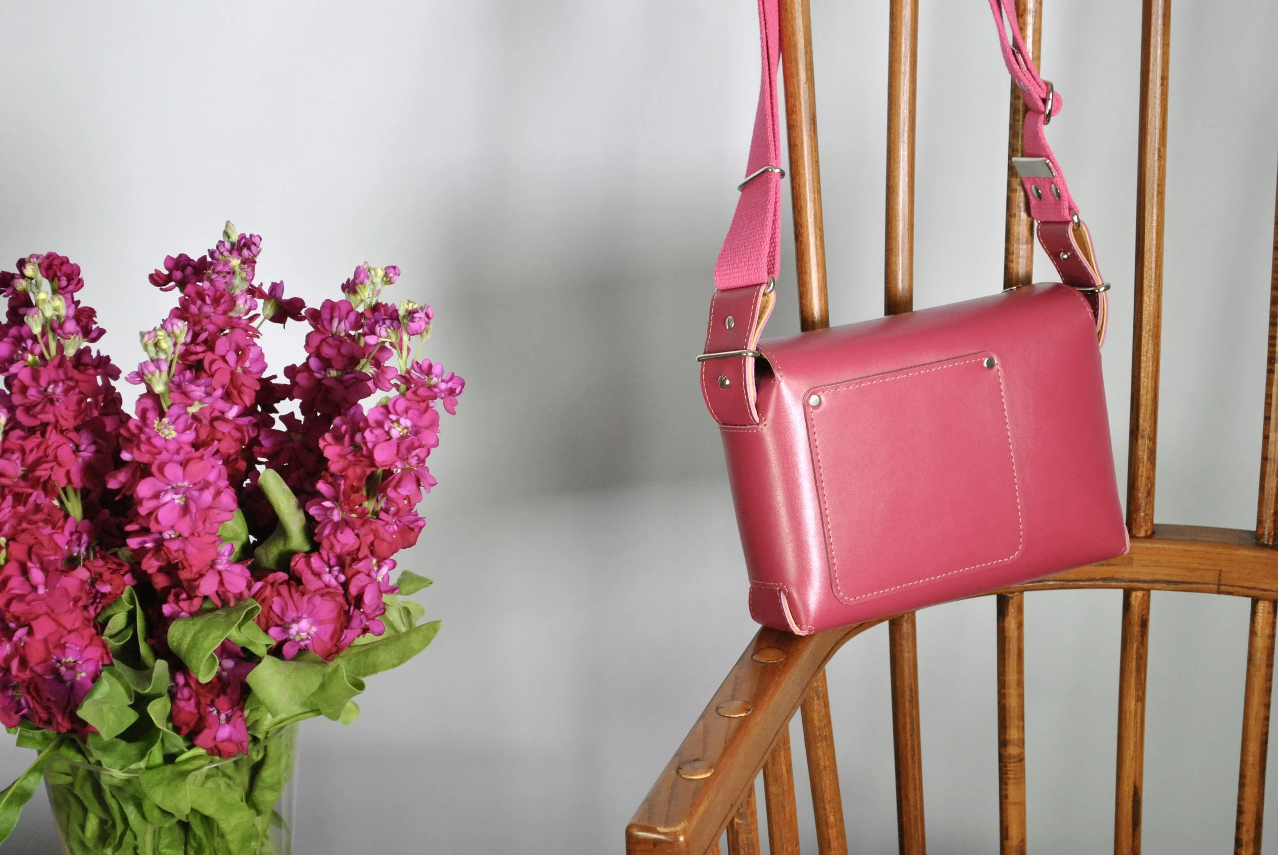 Pink handbag hanging on a wooden chair next to a bunch of pink flowers in a green vase.