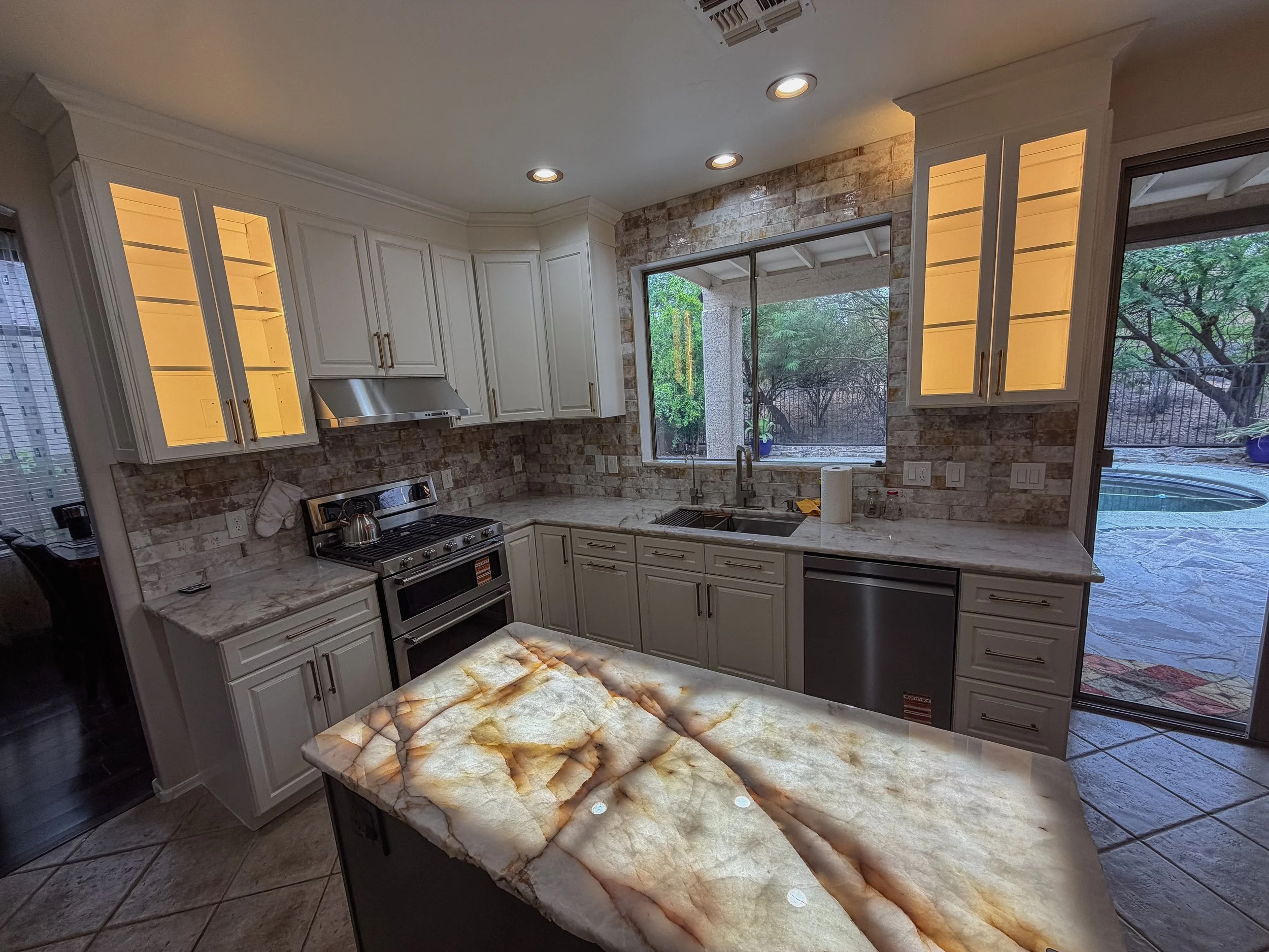 A kitchen with white cabinets, a marble countertop with a matching island, brick backsplash, stainless steel stove, and large windows showing a backyard with trees.