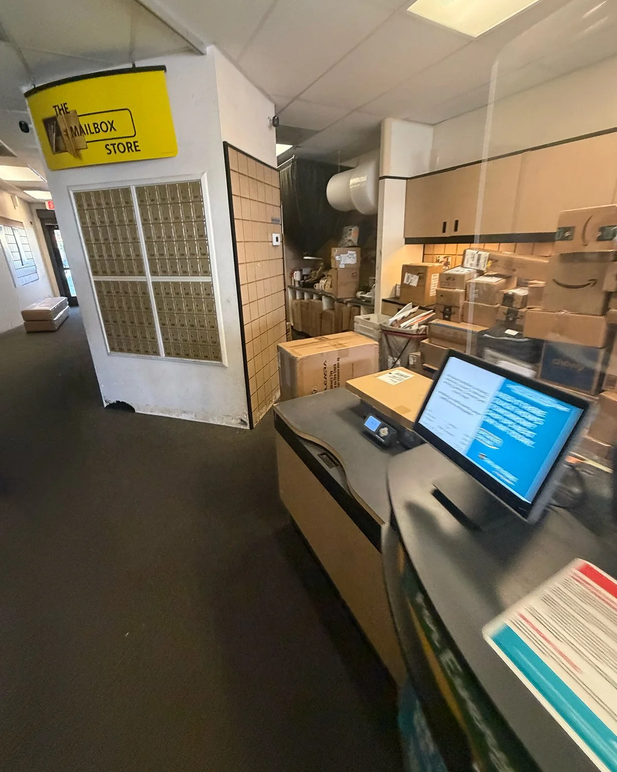Inside a mail store, there are numerous stacked boxes and packages, a checkout counter with a monitor and phone, and a yellow mailbox sign on the wall.