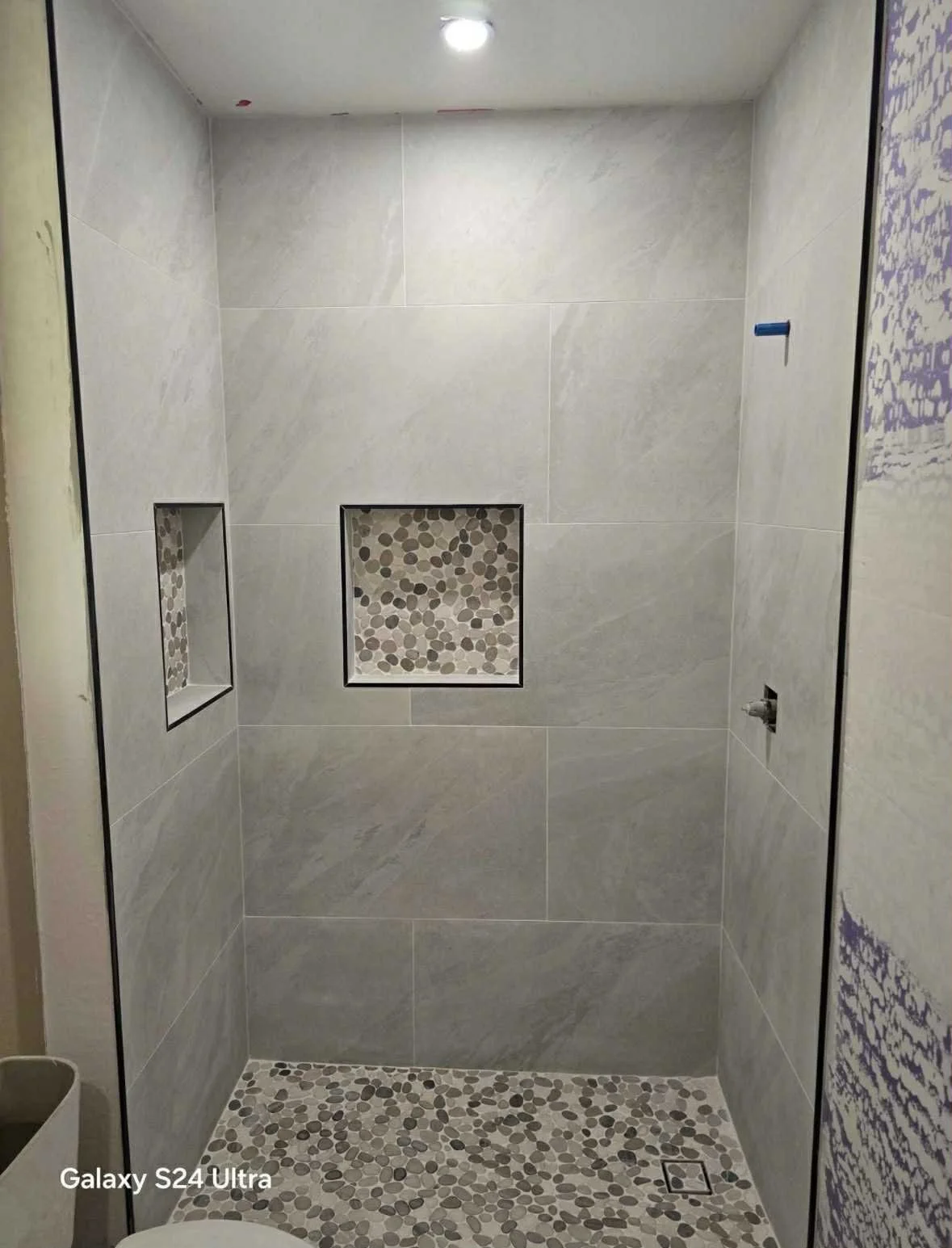 A walk-in shower with large gray tiles on the walls, two recessed shelves with pebble mosaic tiles, pebble mosaic tiles on the shower floor, a ceiling light, and a partially visible toilet in the bottom left corner.