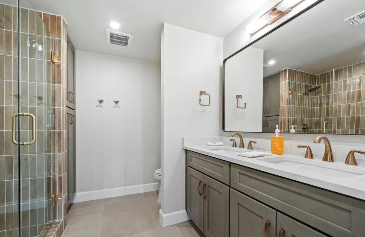 Modern bathroom with double vanity, large mirror, brown faucets, soap dispenser, shower with glass door, and neutral tile decor.