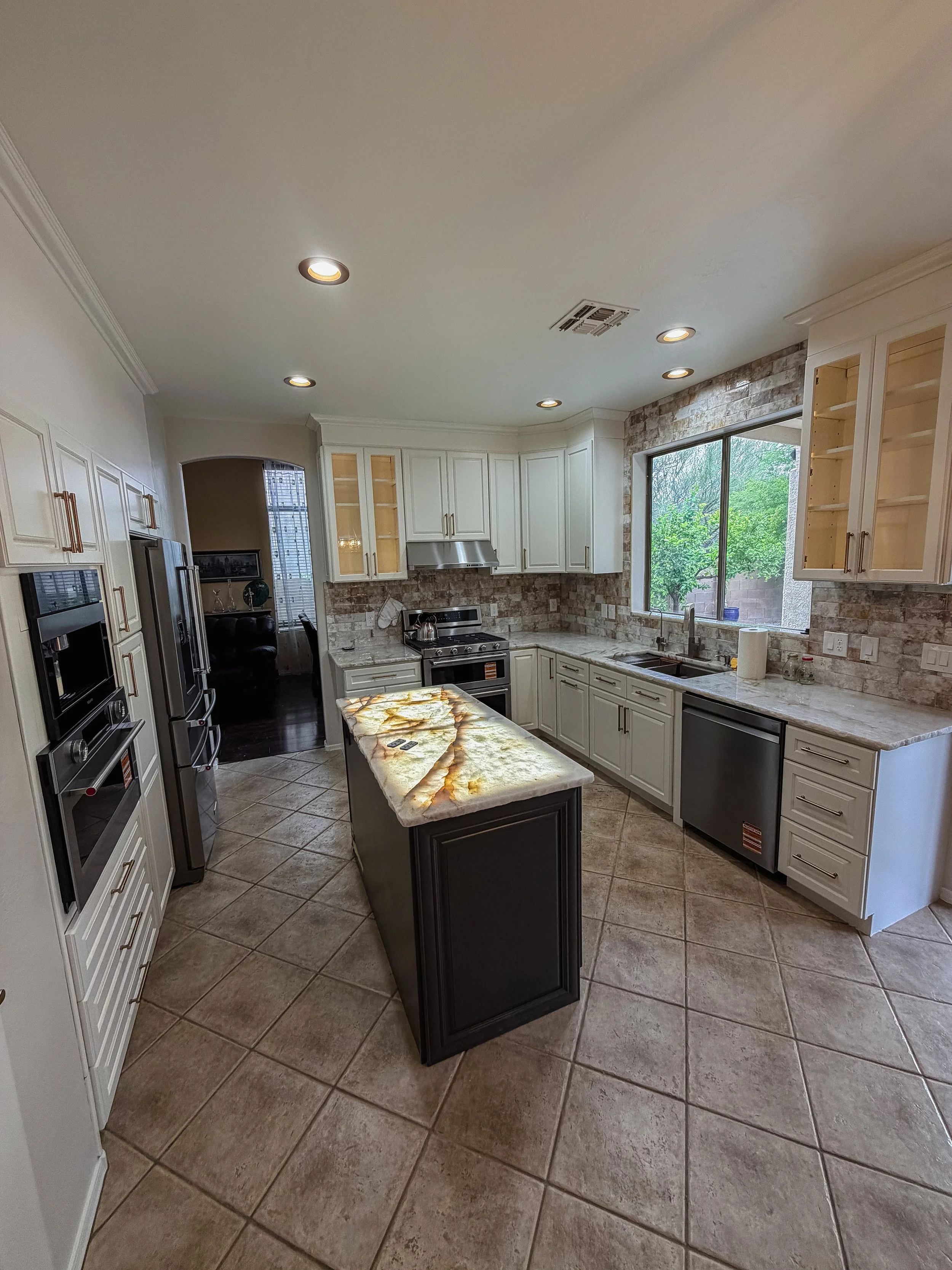 Modern kitchen with white cabinets, stainless steel appliances, granite countertops, a black kitchen island, large window revealing greenery outside, and recessed ceiling lights.