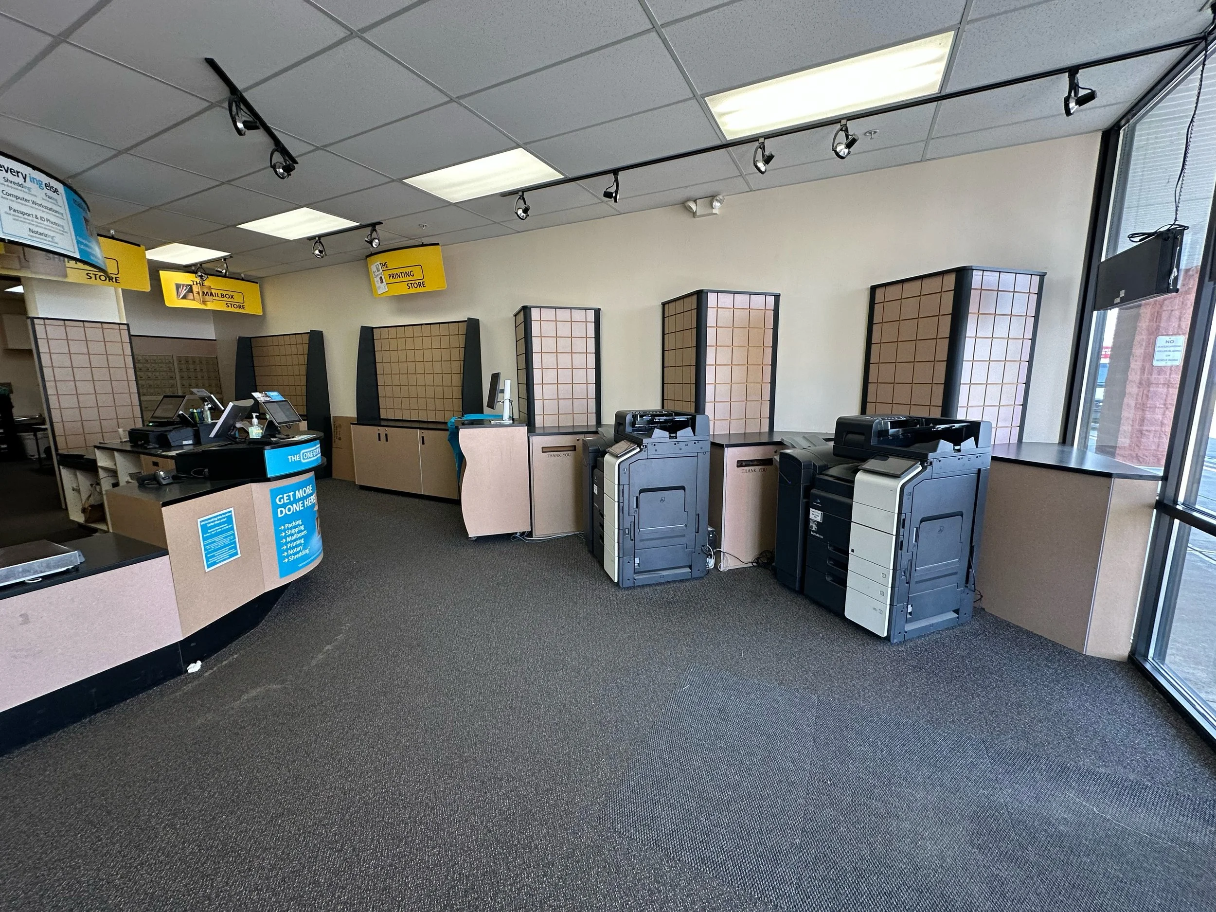 Interior of a retail store or office with service counters, multiple printers, and display panels, near large glass windows with natural light.