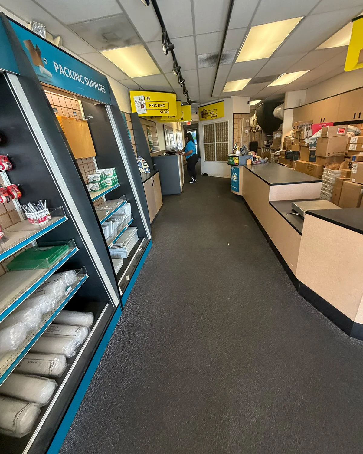 Interior of a retail store or office supply shop with shelves stocked with supplies, a counter, and signages indicating printing and packaging supplies, with a person working near the back.