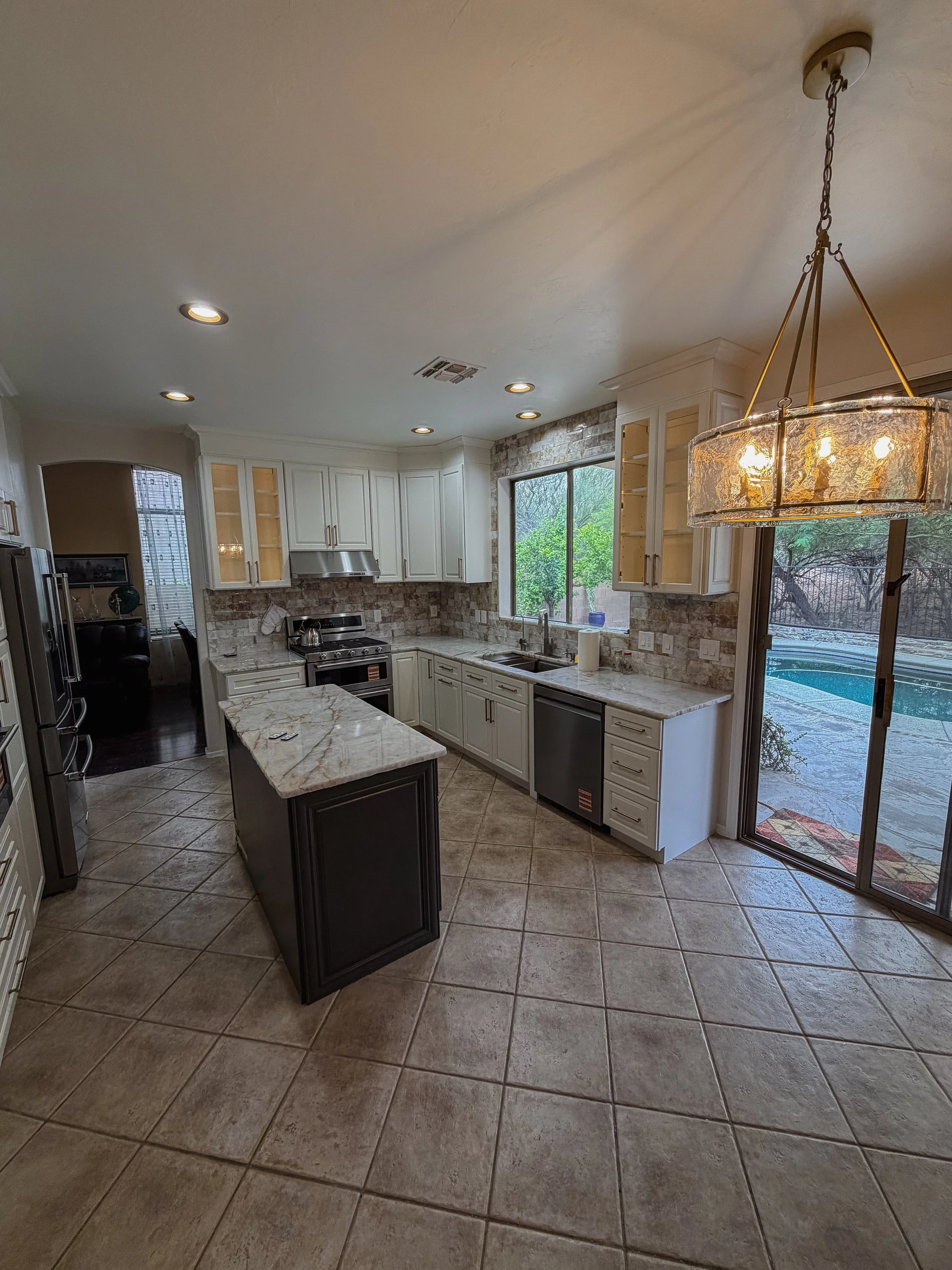 A modern kitchen with white cabinets, a marble countertop, stainless steel appliances, and a tiled floor. There's a large window above the sink, a glass-paneled door leading outside, and a decorative chandelier hanging from the ceiling.