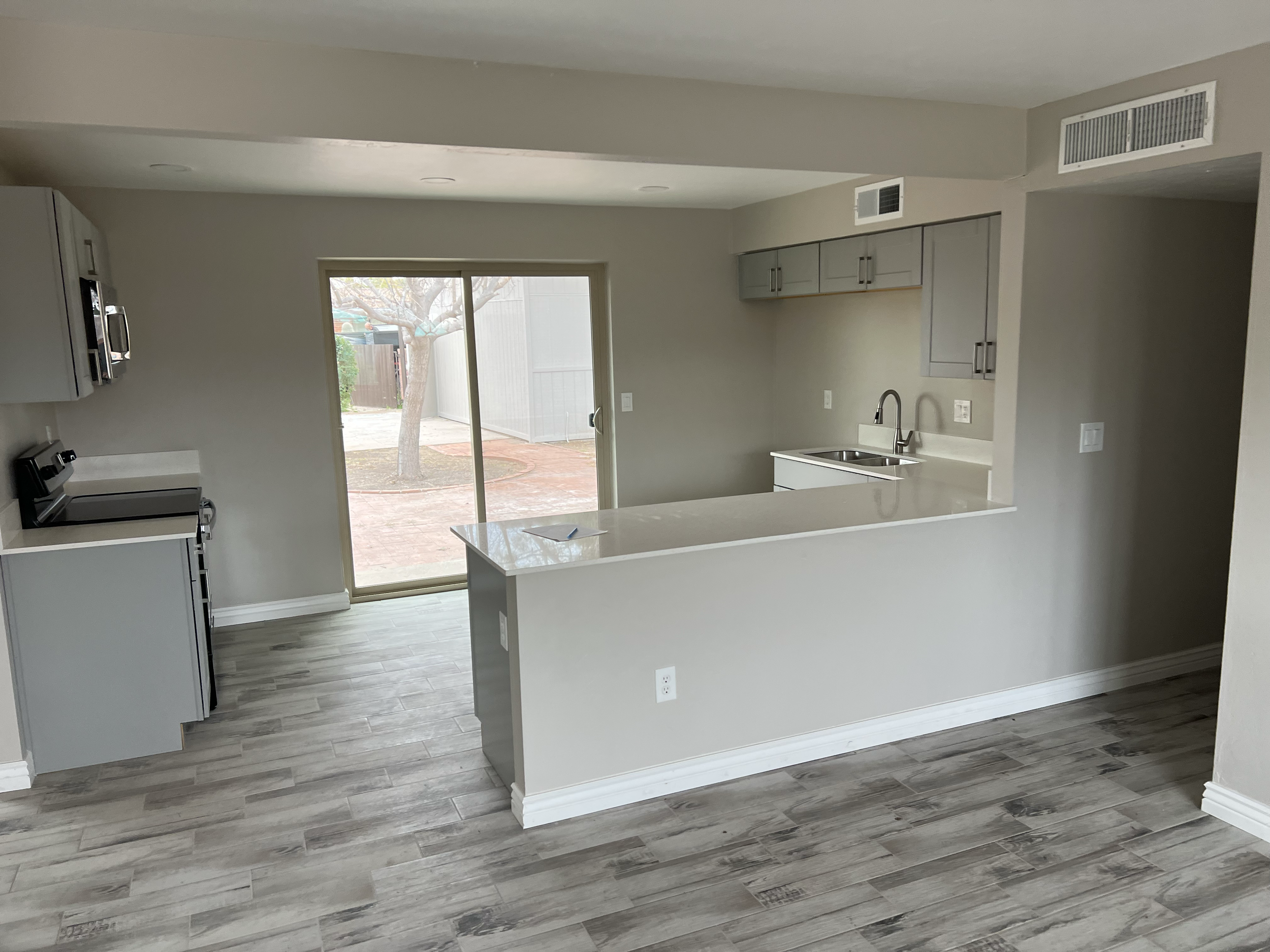 Empty kitchen with white countertops, gray cabinets, a stainless steel sink, a stove, and a sliding glass door leading to the backyard with a tree.