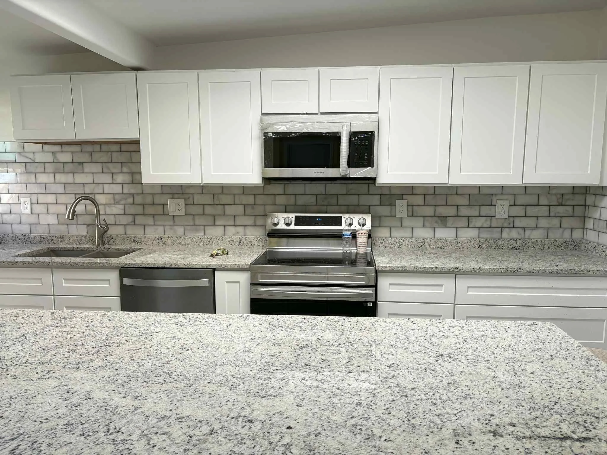 A modern kitchen with white cabinets, granite countertops, gray brick backsplash, stainless steel microwave above stove, stainless steel oven, dish soap, a water bottle, and a paper cup near the stove, and a double sink with faucet.