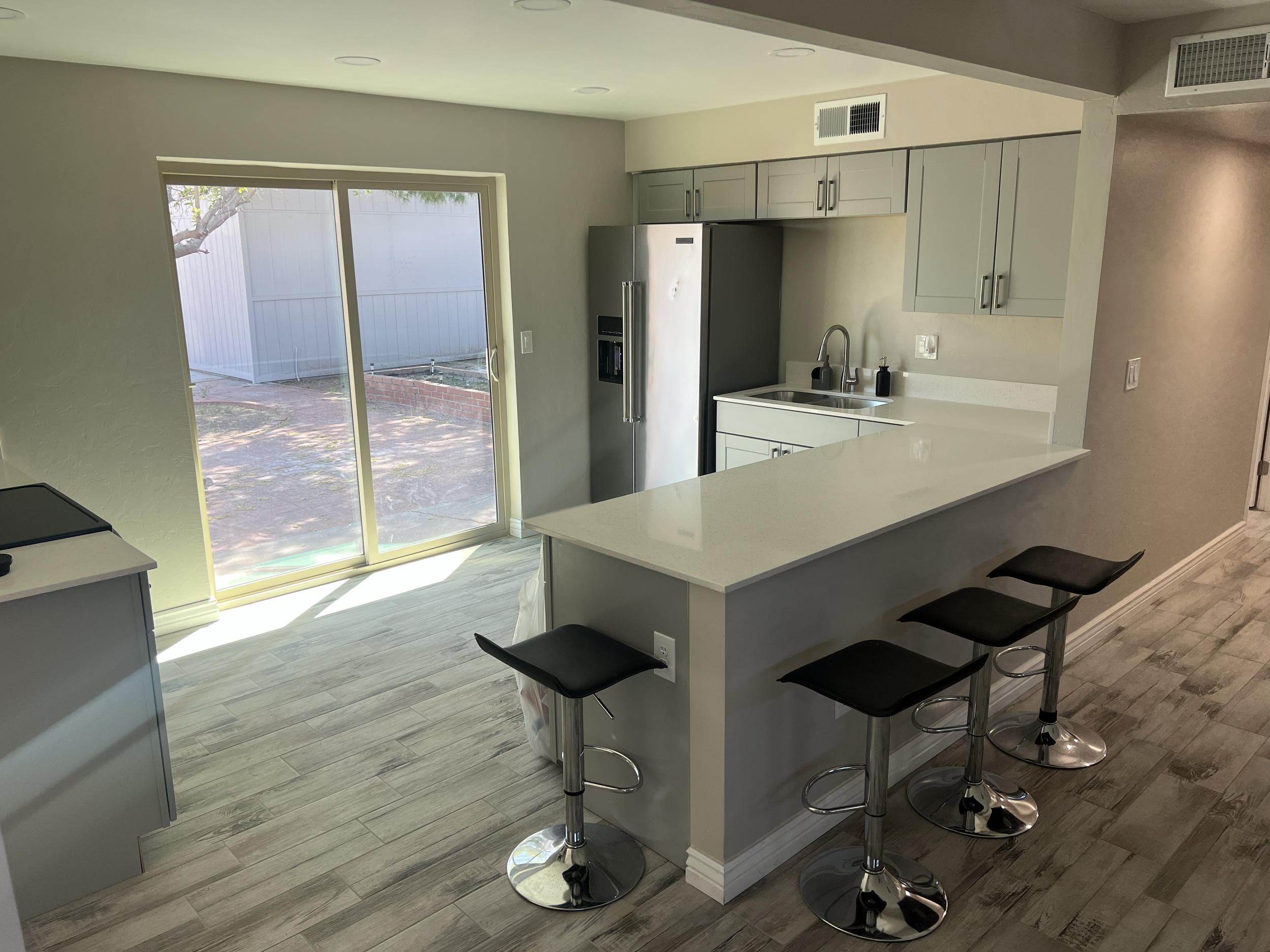 A modern kitchen with gray cabinets, a stainless steel refrigerator, and a white island with black barstools. There is a sliding glass door leading to the backyard, with a view of a tree and a white fence.