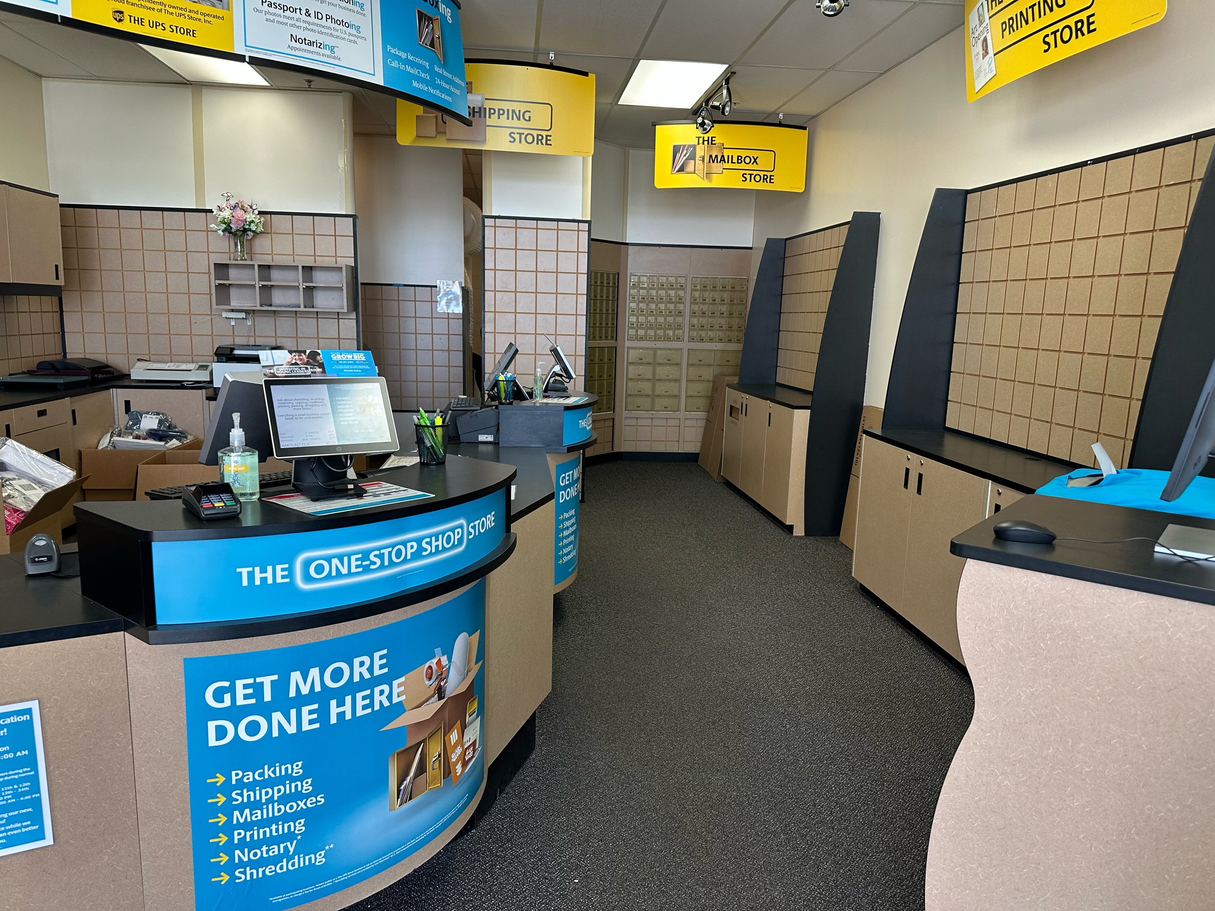 Inside a USPS store with yellow signs hanging from the ceiling indicating sections like the Mailbox Store, Shipping Store, and Printing Store. The counter area has various packing and mailing supplies, computers, and signage promoting services like p