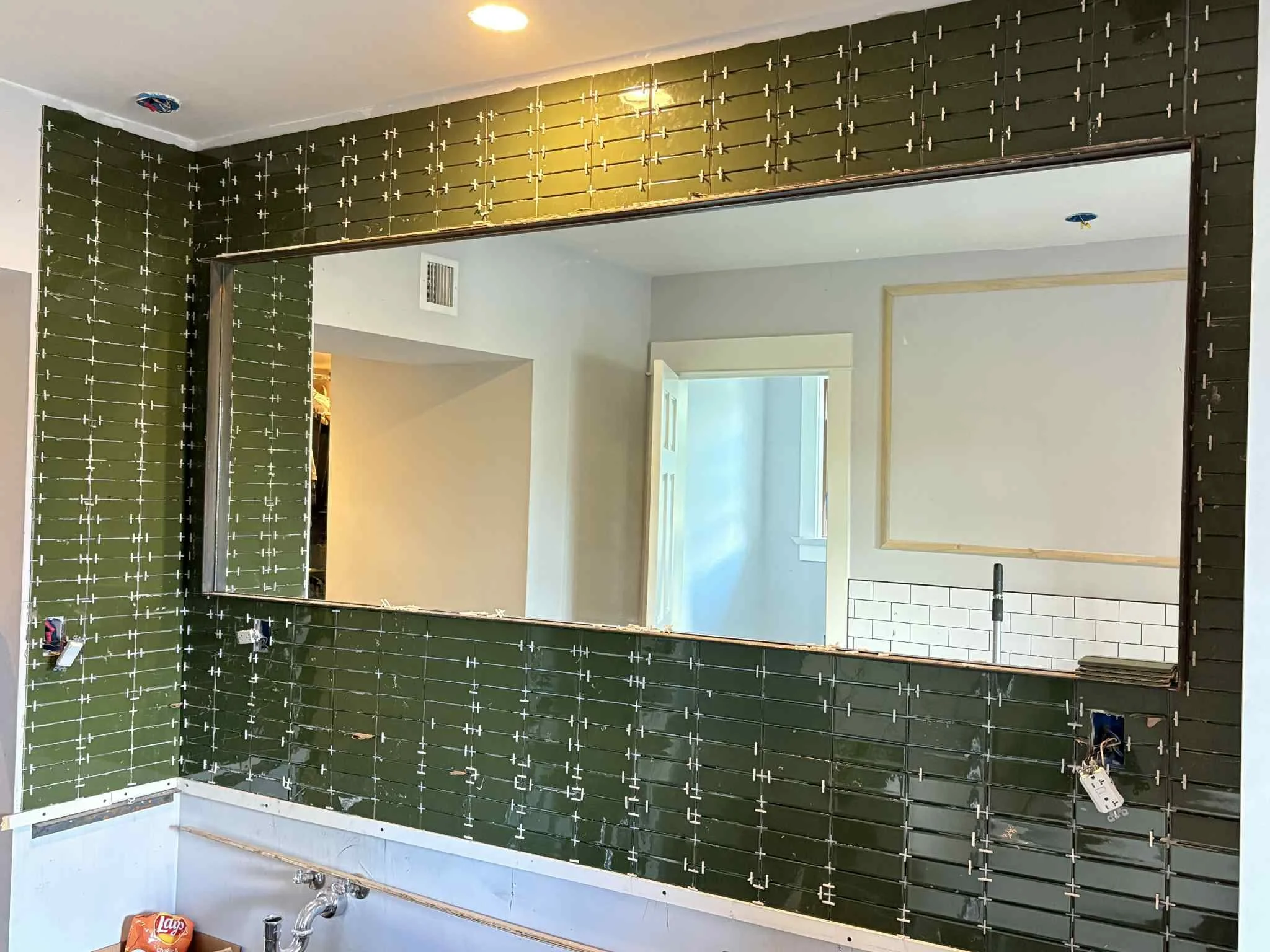 A large mirror reflecting a doorway and partial interior room, mounted on a wall with green tile or paneling showing white tile spacers and exposed wiring, indicating ongoing construction or renovation.
