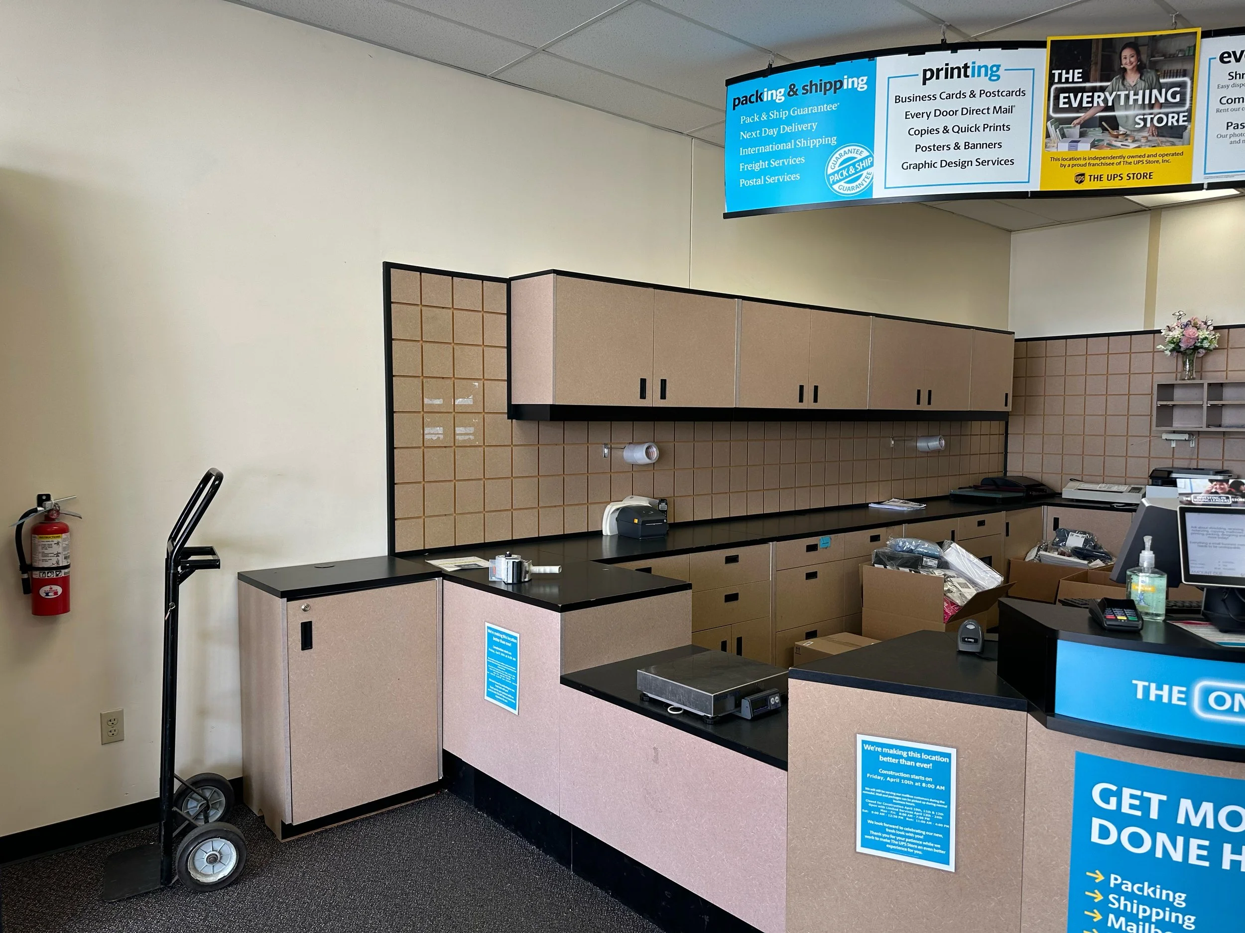 Interior of a store counter area with beige cabinets, a black countertop, a cash register, and signs advertising shipping and printing services. There is a fire extinguisher on the wall, a hand truck, and a small flower arrangement on the side. A blu