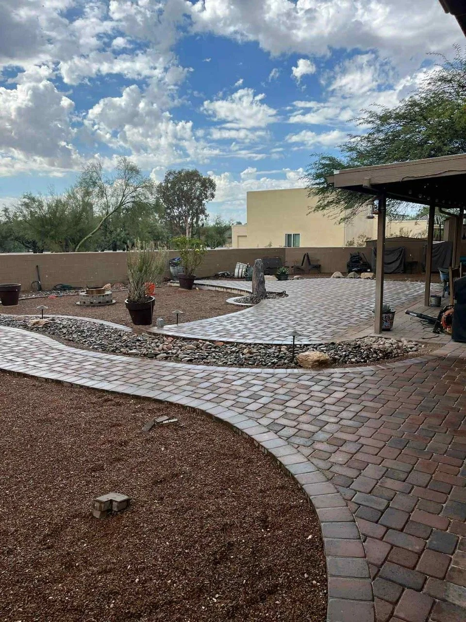 Backyard with newly paved brick walkway, decorative rocks, a few potted plants, and a covered patio area. Trees and a blue sky with scattered clouds in the background.